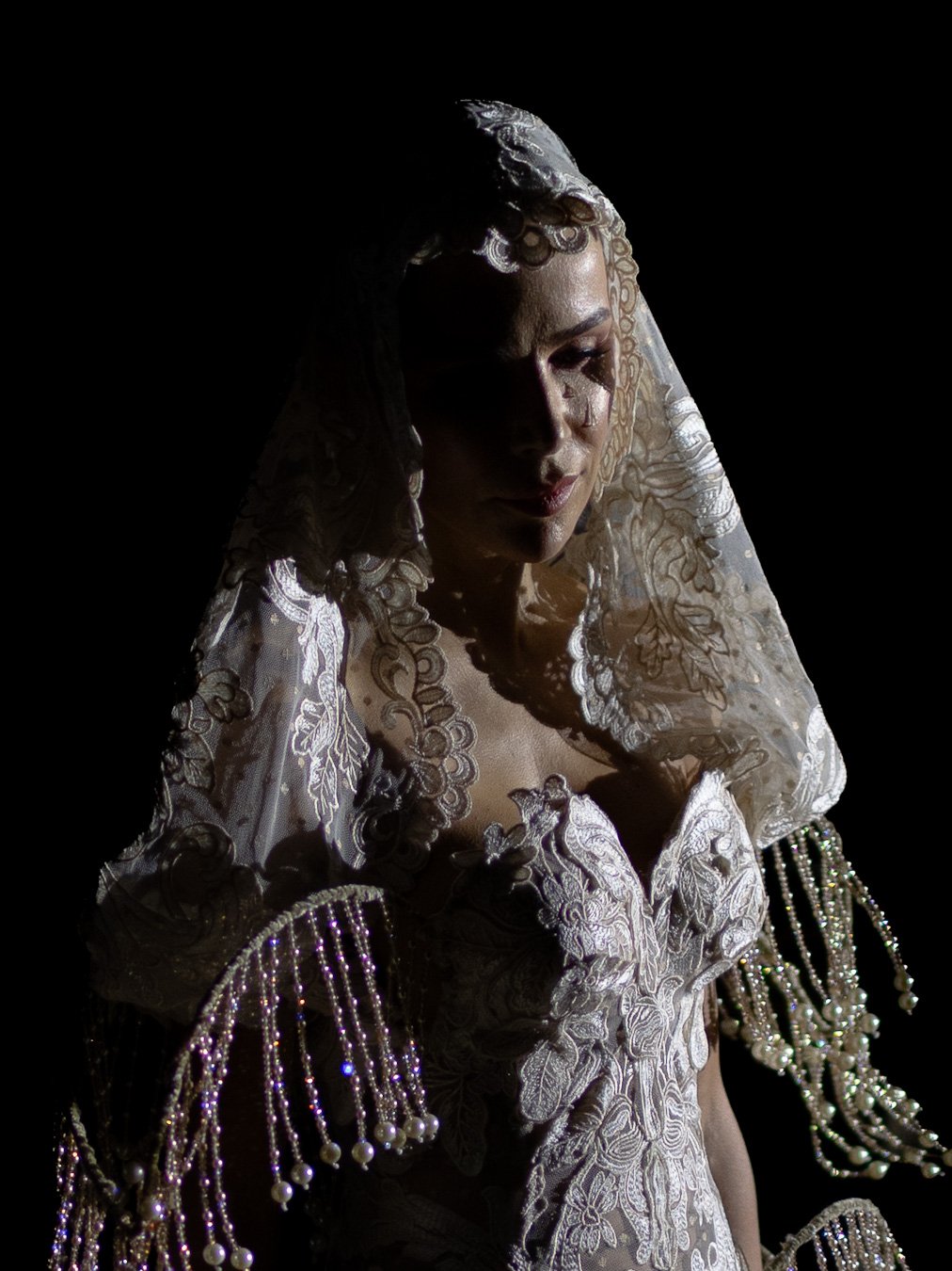 A woman wearing ornate lace bridal gown with pearl and bead embellishments, service in a dark setting with lighting highlighting her face.
