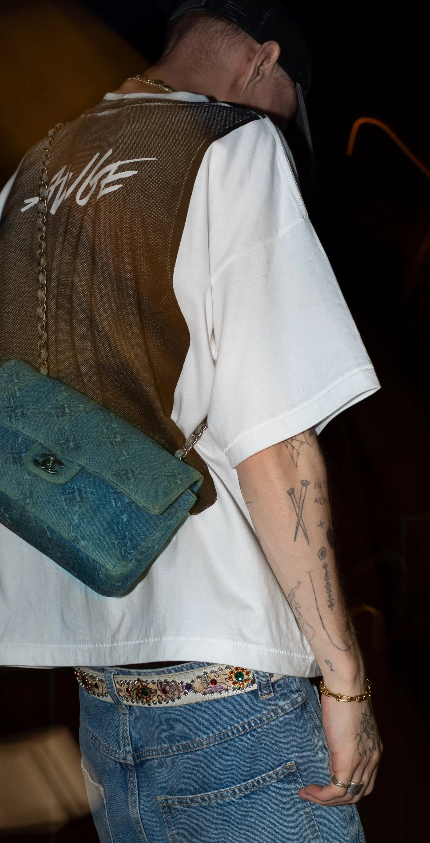 A person wearing a white t-shirt with tattoos on the arm, a chain necklace, a blue designer purse with a chain strap, and embellished jeans with a decorative belt, standing in a dark environment.