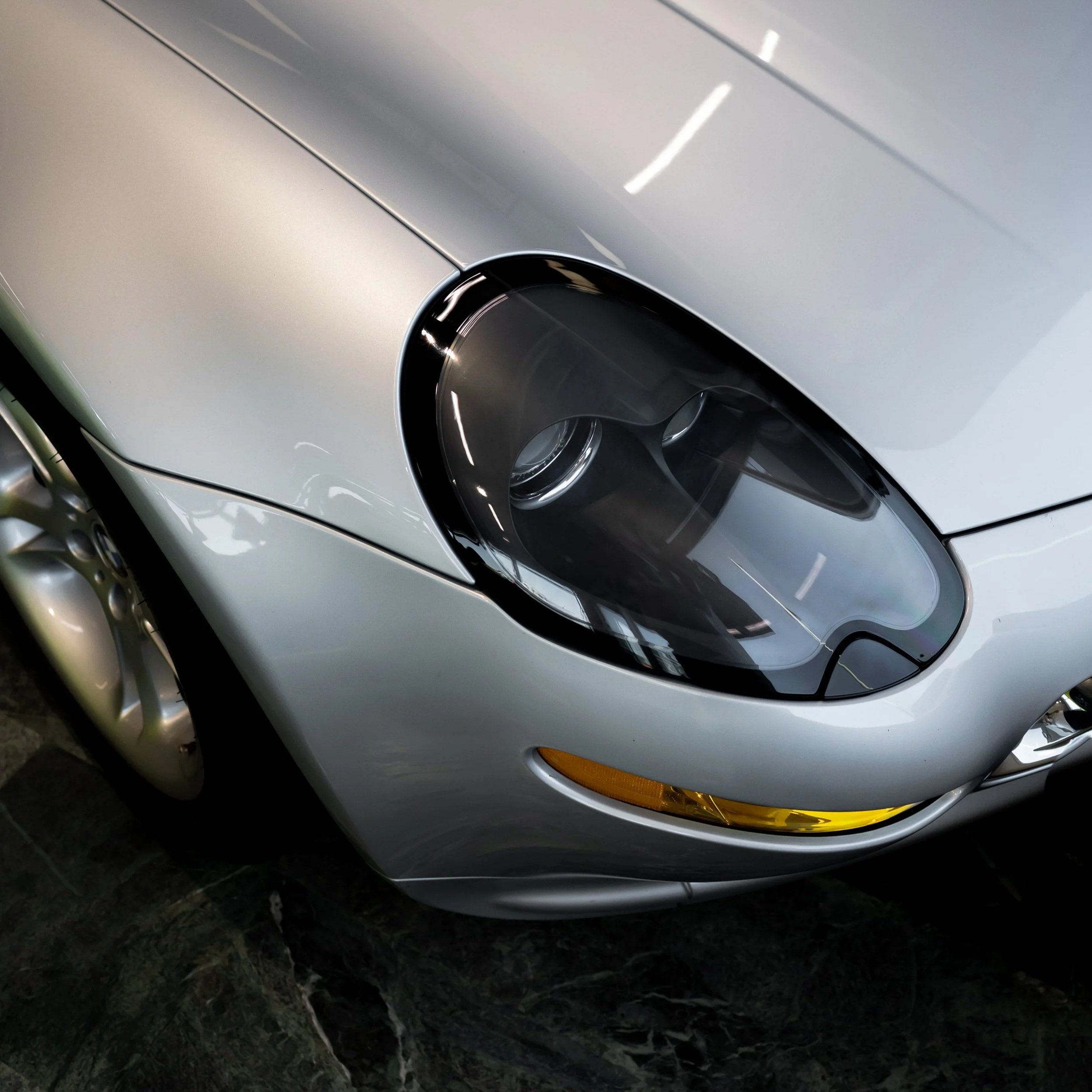 Close-up of the front part of a silver sports car, showing the headlight and wheel, parked on a grassy and rocky surface.
