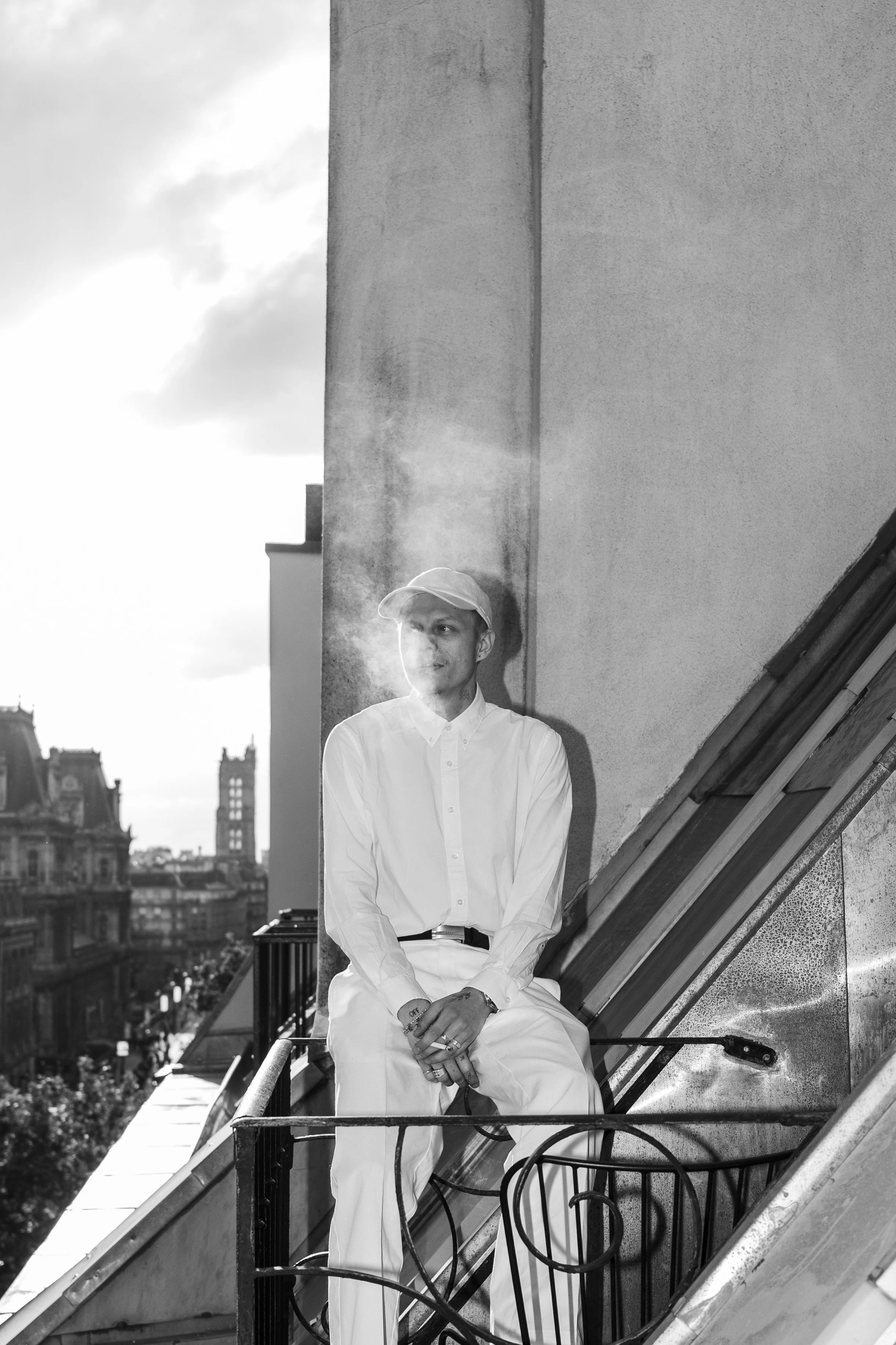 A young man in white clothing and a cap is sitting on a rooftop ledge, holding a cigarette, with city rooftops and buildings in the background. Sunlight is shining, creating a smoking trail in the air.