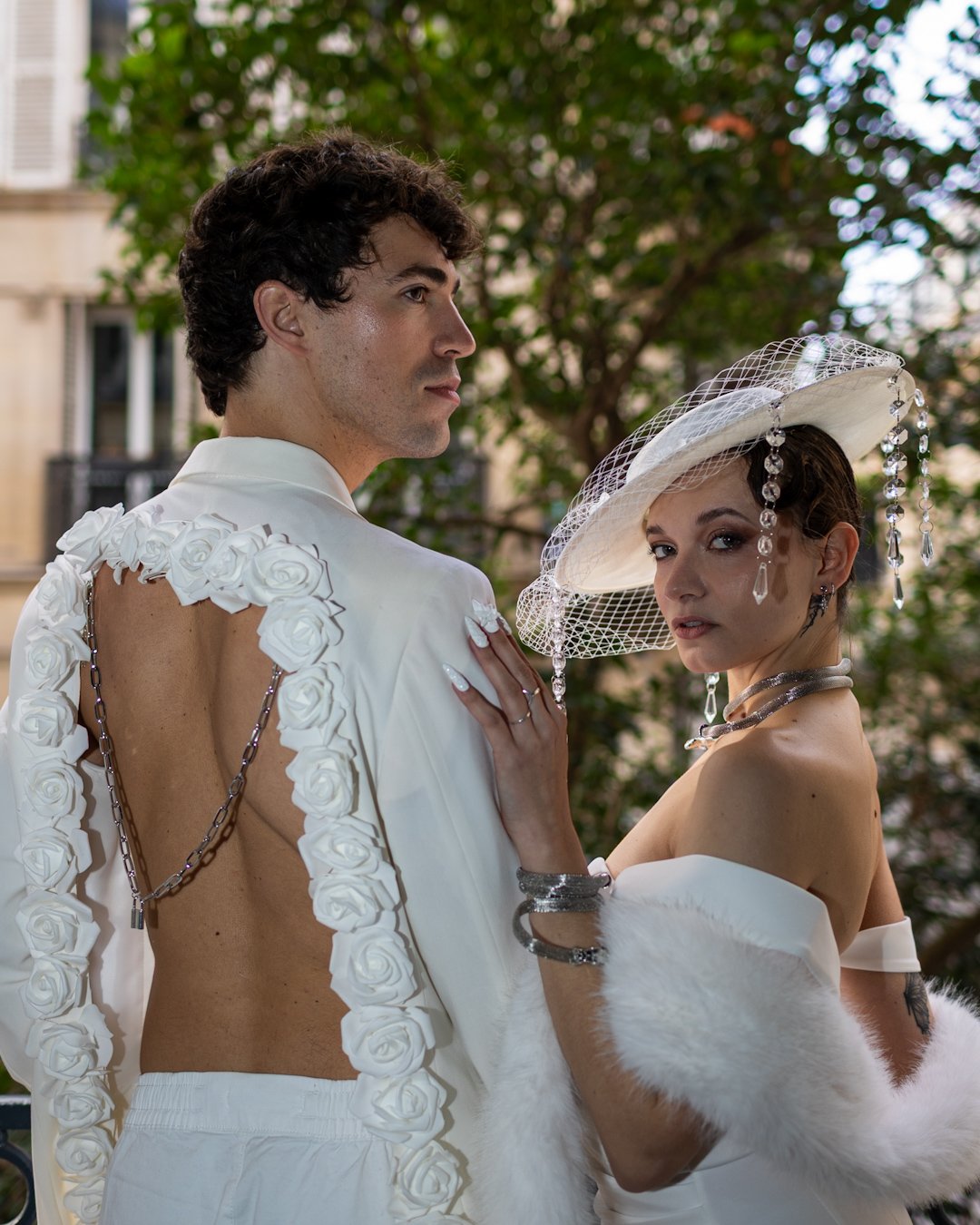 A man and woman dressed in luxury fashion, posing outdoors with trees and buildings in the background. The man wears an open-back white shirt decorated with roses and chain details. The woman wears a large white hat with pearls and netting, jewelry, and a white fur stole.