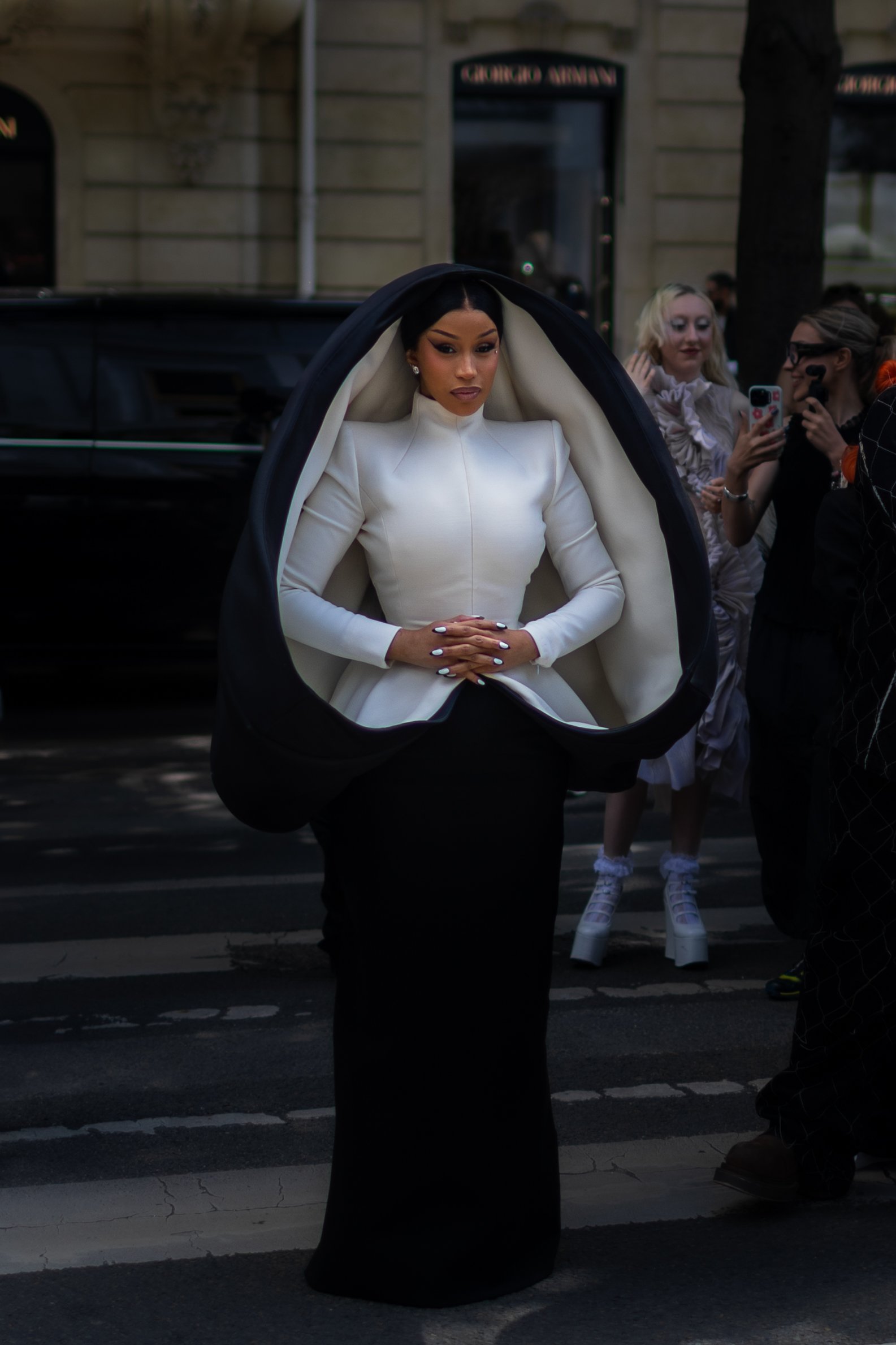 A woman with dark hair in a white dress with structured shoulders, seated inside a black and white garment resembling a large circular cape or hood, is standing on a city street during daytime, with two women to the right taking a photo and smiling.