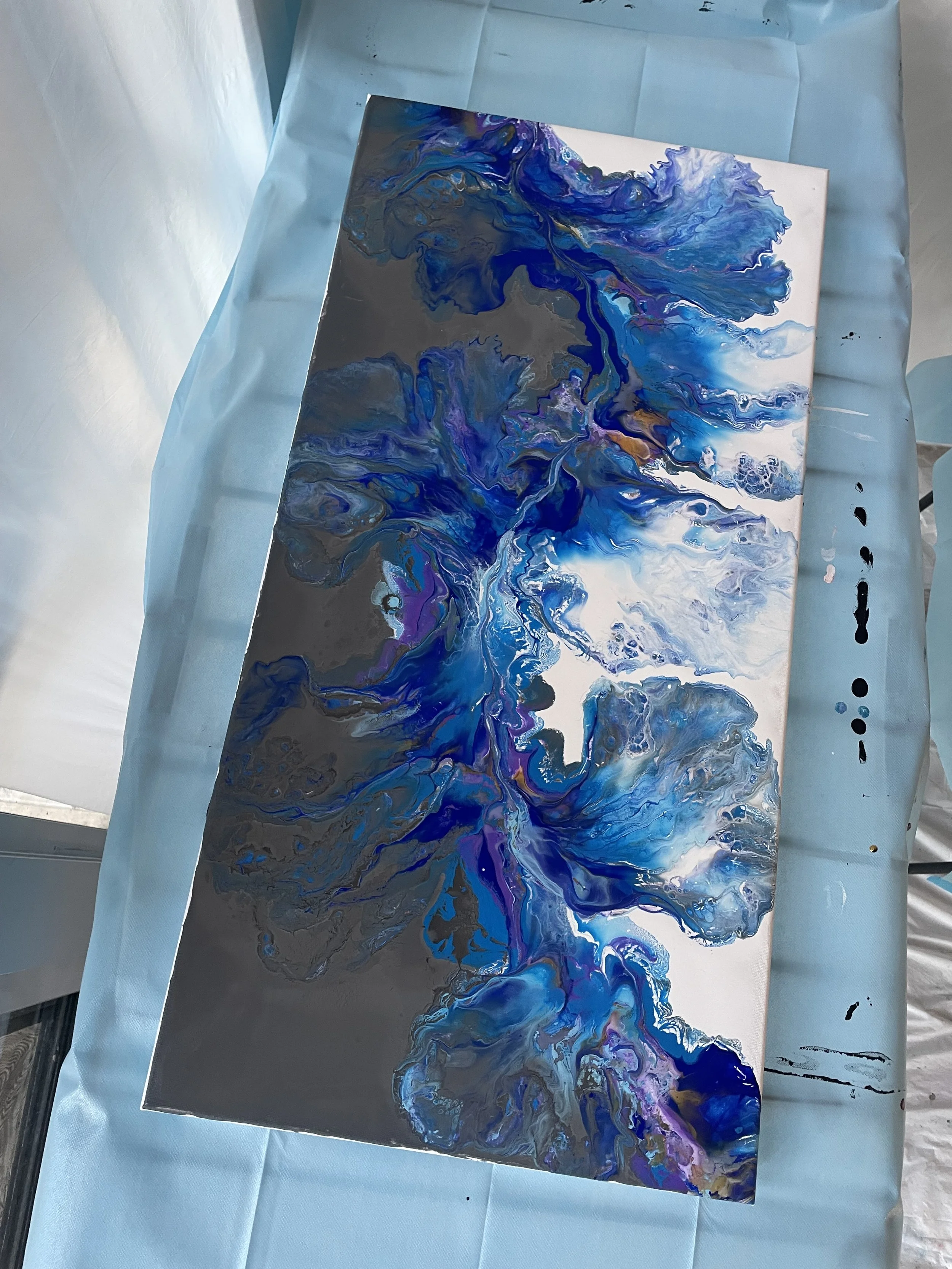 A canvas with an abstract fluid art painting featuring swirling shades of blue, purple, black, and hints of gold, resting on a light blue protective sheet.