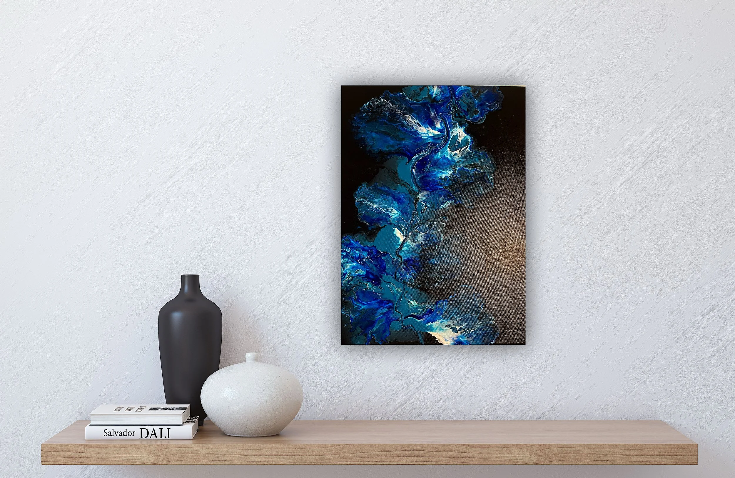 Decorative abstract blue and black watercolor painting hanging on a white wall above a wooden shelf. On the shelf, there's a black vase, a white vase, and stacked books with a black-and-white color theme.