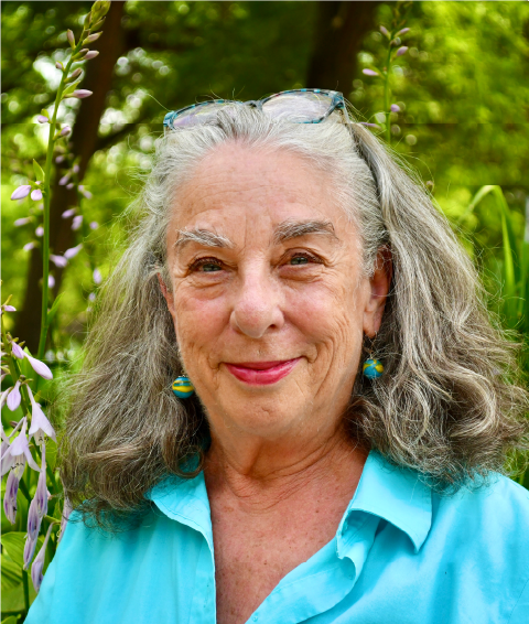 A smiling elderly woman with gray hair, wearing glasses on her head, a turquoise shirt, and colorful earrings, standing outdoors in front of green trees and purple flowers.