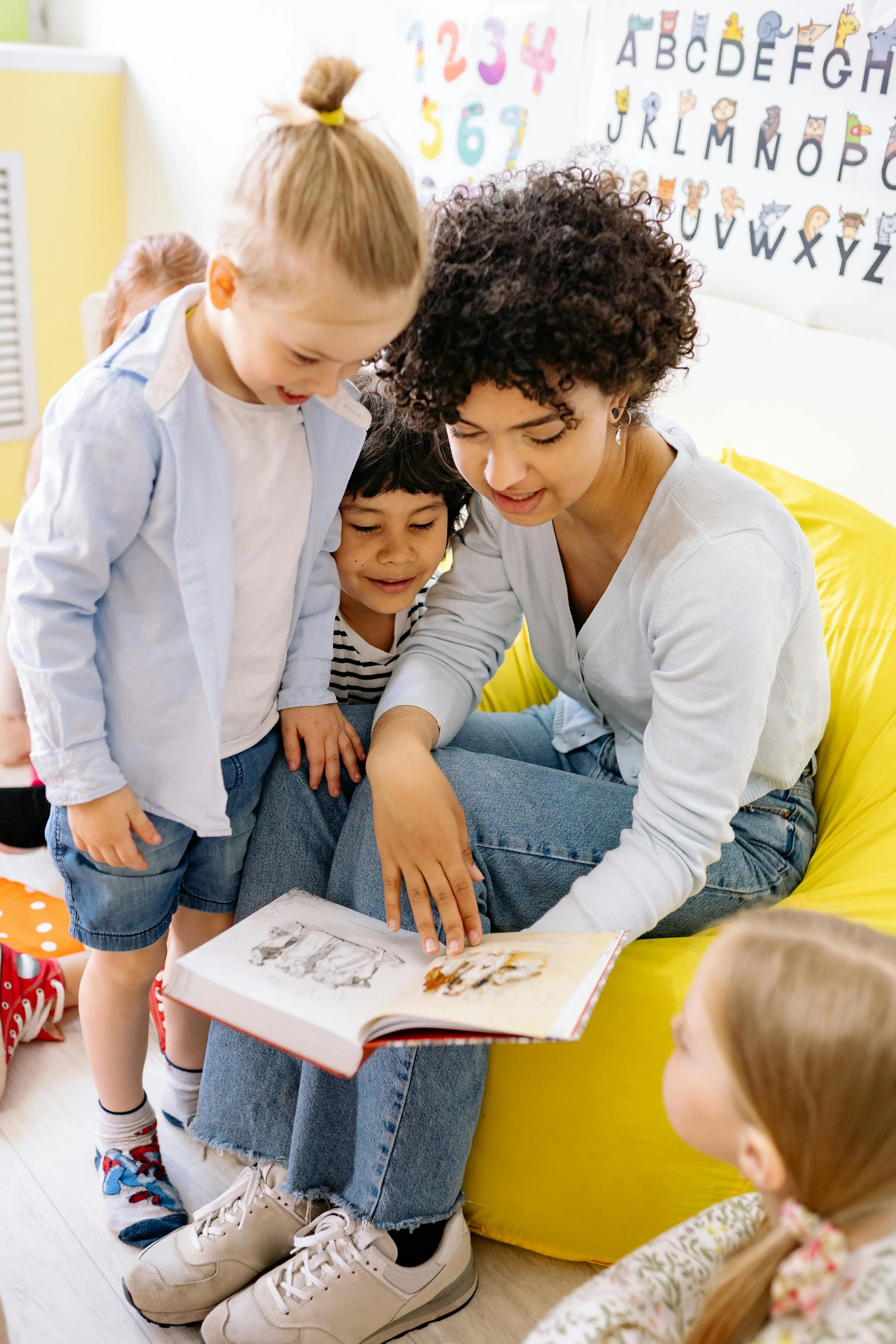A teacher reading a picture book to a group of younger children in a classroom decorated with colorful numbers and alphabet chart on the wall.