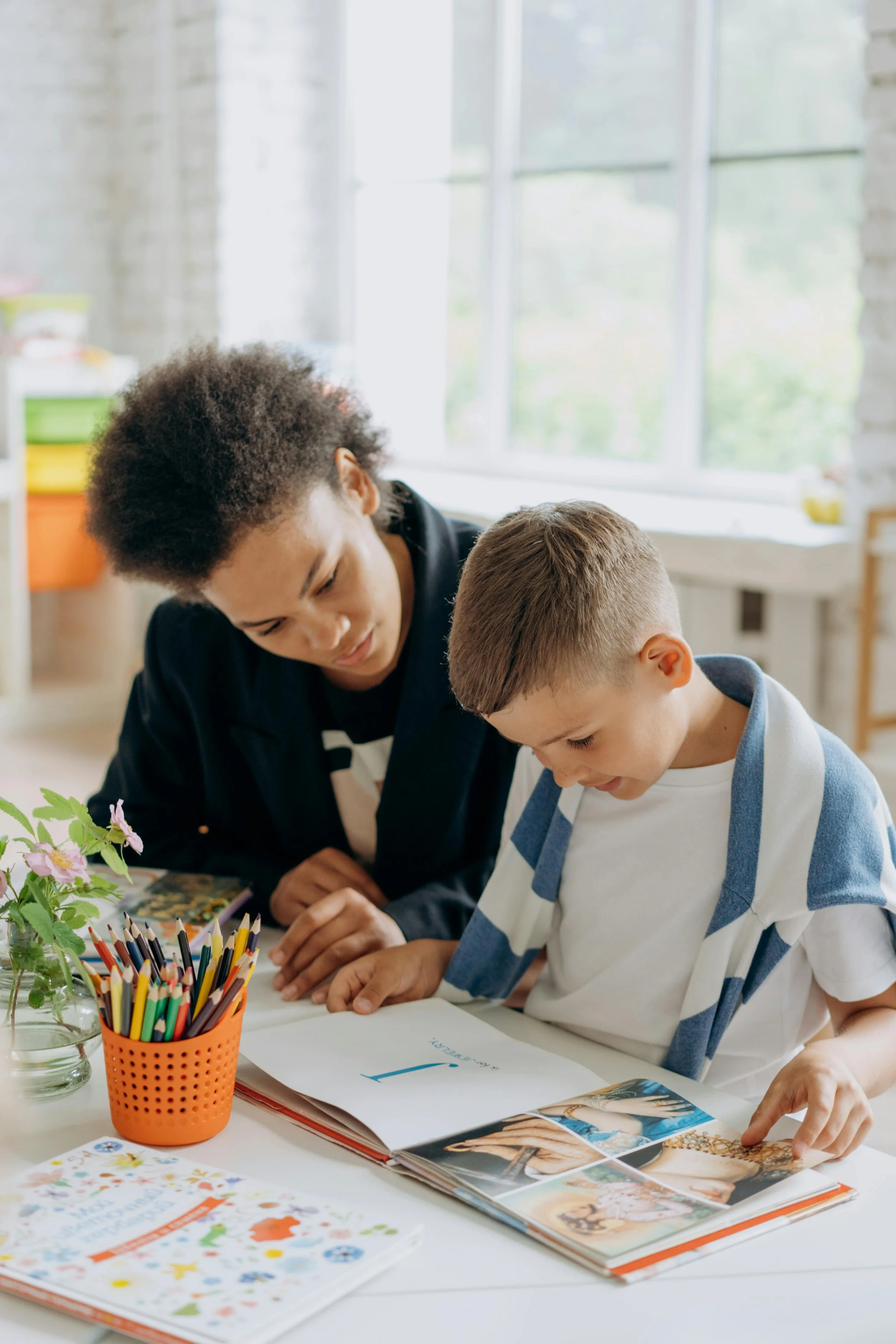 A younger boy with glasses and a striped shirt is learning about art with a woman, possibly his teacher, in a bright classroom filled with art supplies and books.
