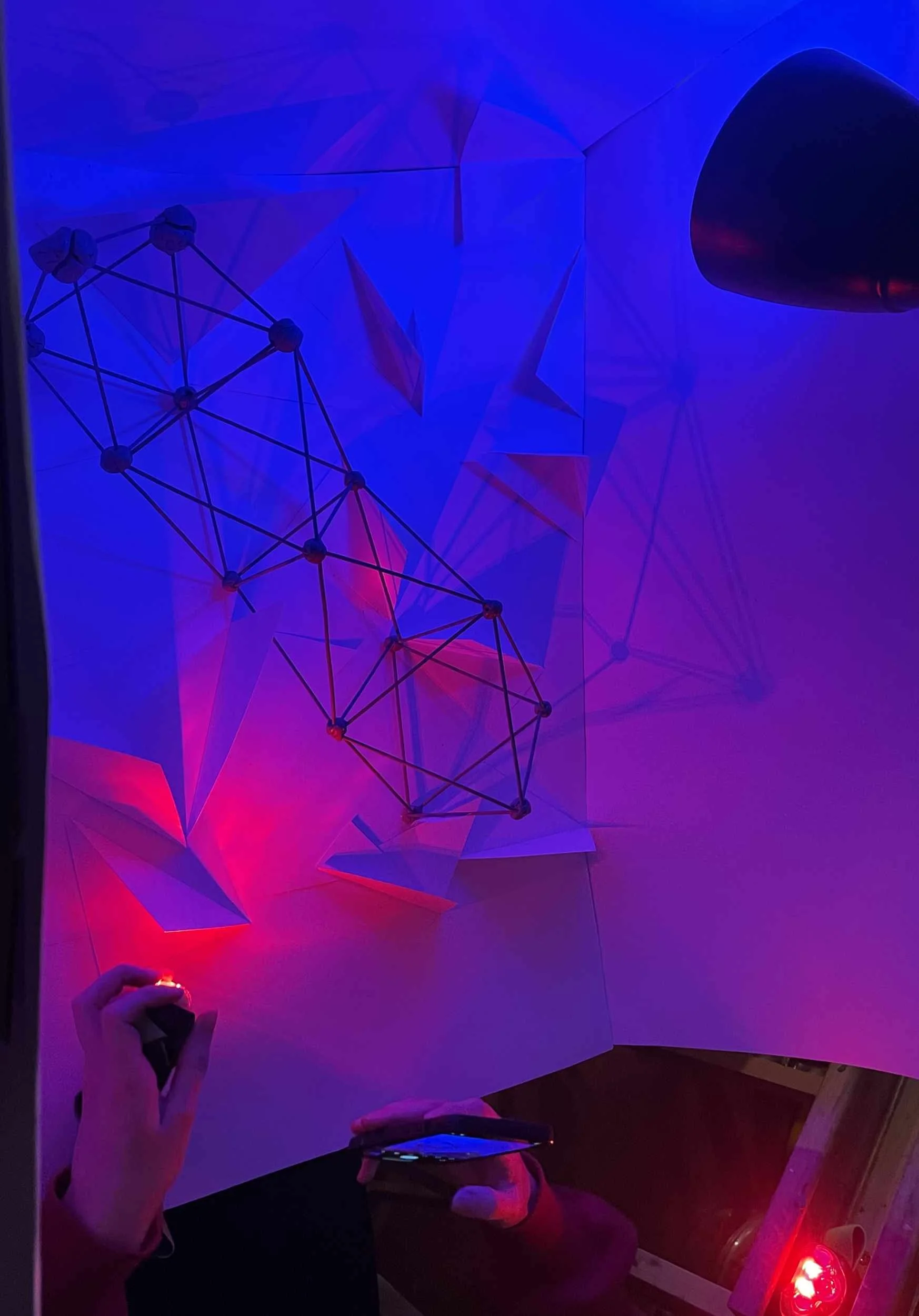 Model oświetlony światłem czerwonym i niebieskim naraz. Makieta próbnej scenografii.  Atmosfera scenografii tworzona światłem i silną geometrią. Prętowa struktura leży otoczona plaszczyznowymi trójkątami.