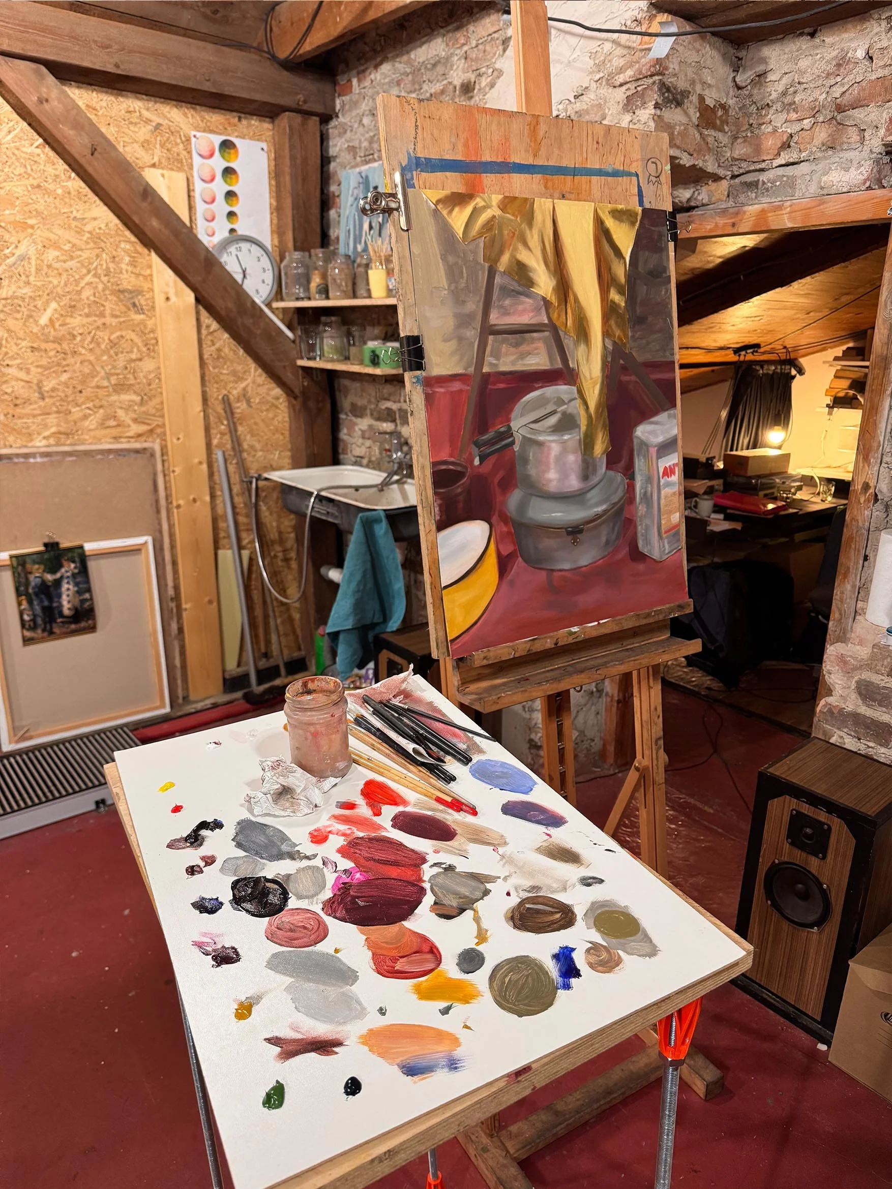 An artist's workspace with a palette filled with various paint colors, a jar of water, and paintbrushes on a table. In the background, there is a painting of a kitchen scene with pots, a clock, shelves with jars, and walls with brick and wooden panel