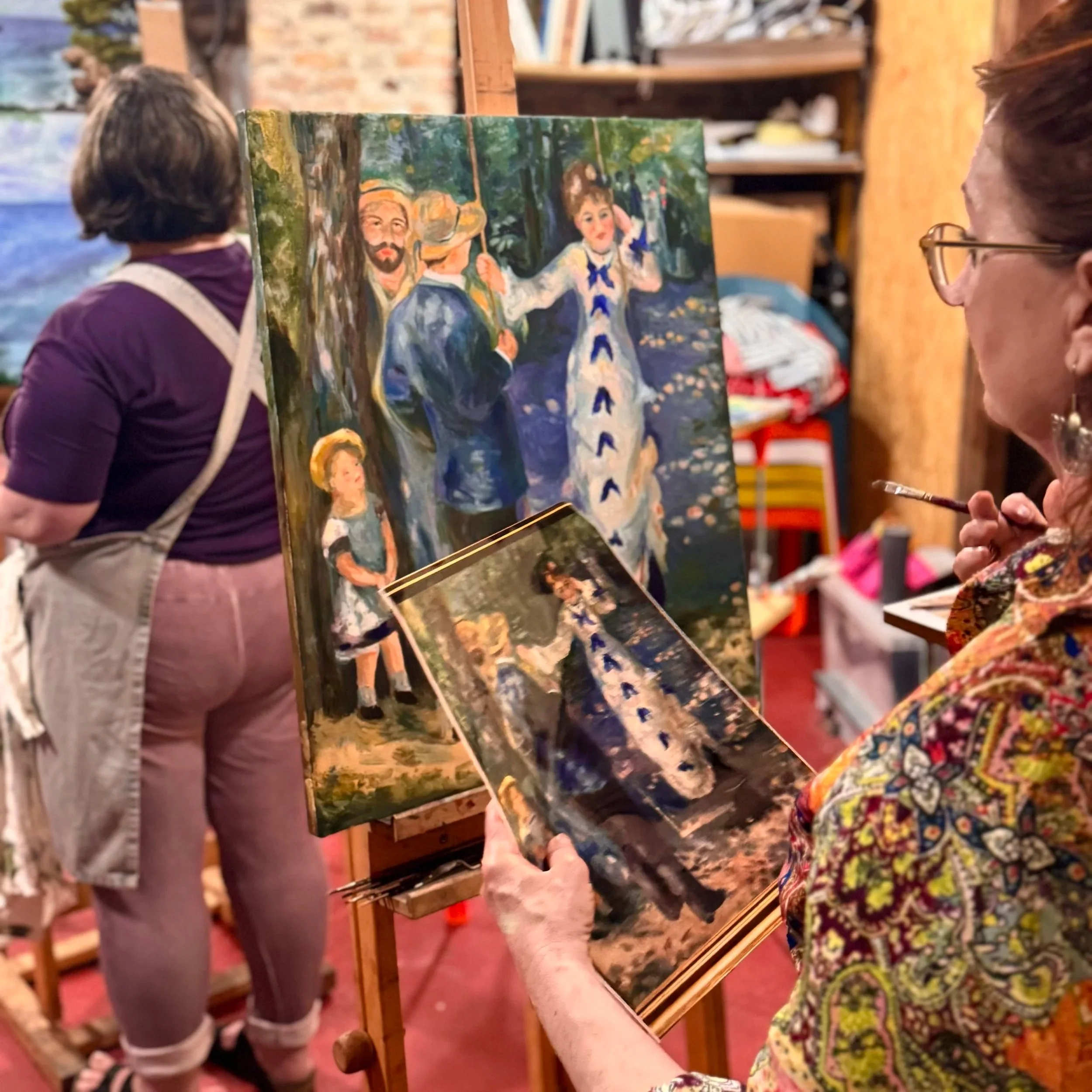Kursantka maluje farbami olejnymi kopie obrazu Chuśtawka (Auguste Renoir 1876) w prawym ręku trzyma pędzel w lewym fotografię oryginału. Na sztalugach jest jej obraz. W tle widać inna malującą osobę i pracownie.