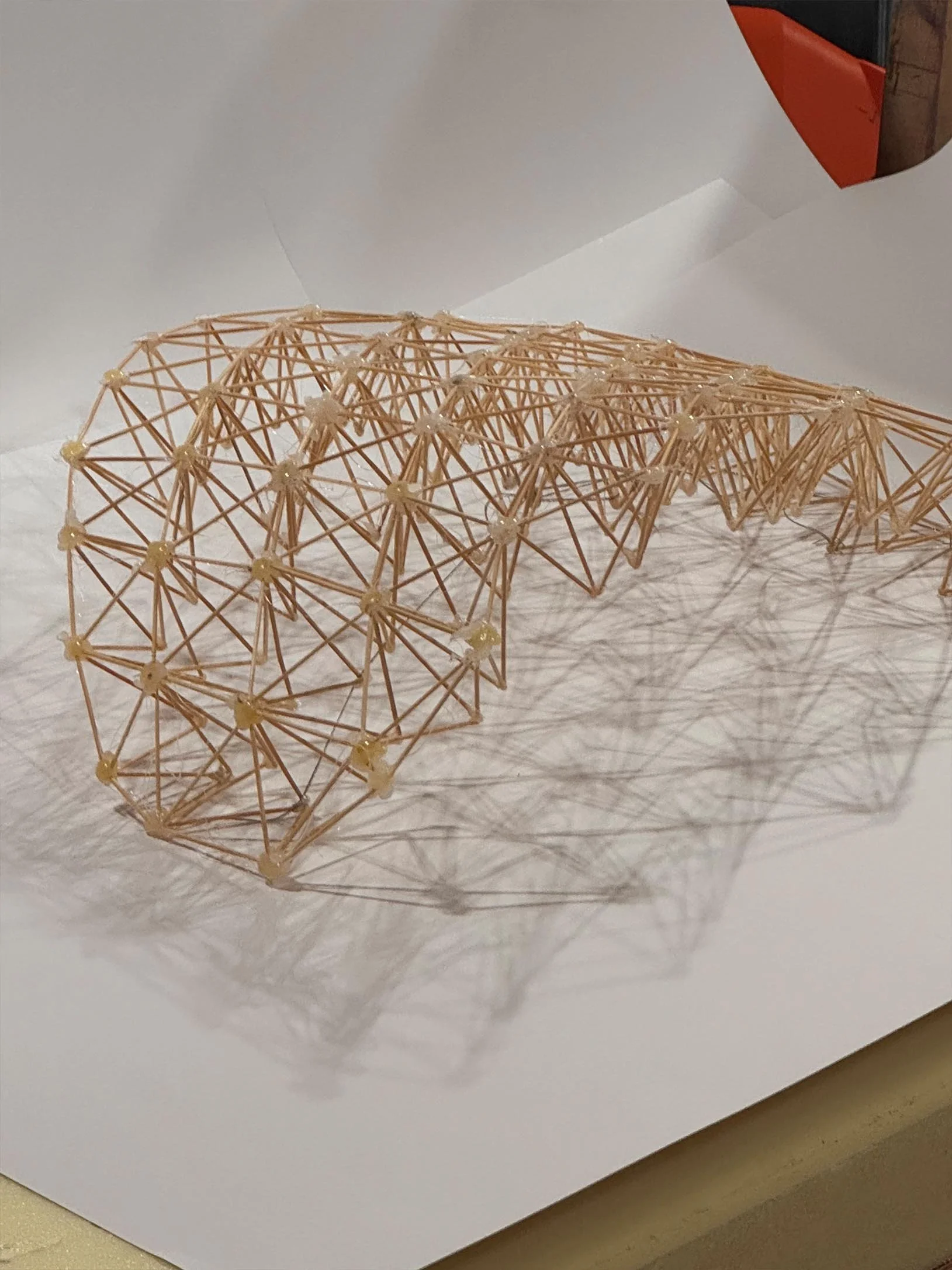 A geometric sculpture made of thin wooden sticks and small white connectors, placed on a white paper-covered surface.