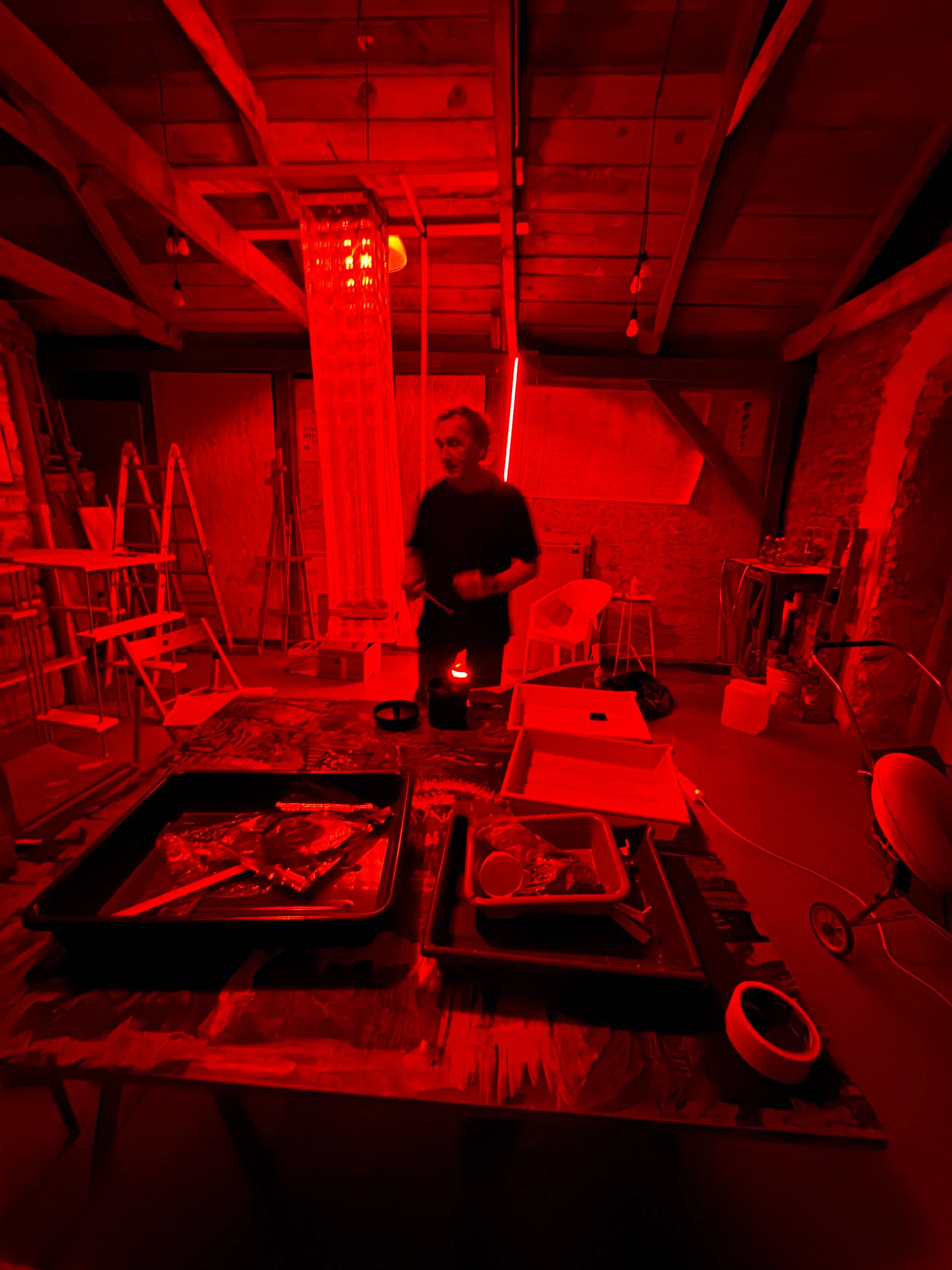 A person standing in a room bathed in red lighting. The room contains ladders, chairs, and various supplies and equipment. The ceiling is wooden, and there are brick walls visible.
