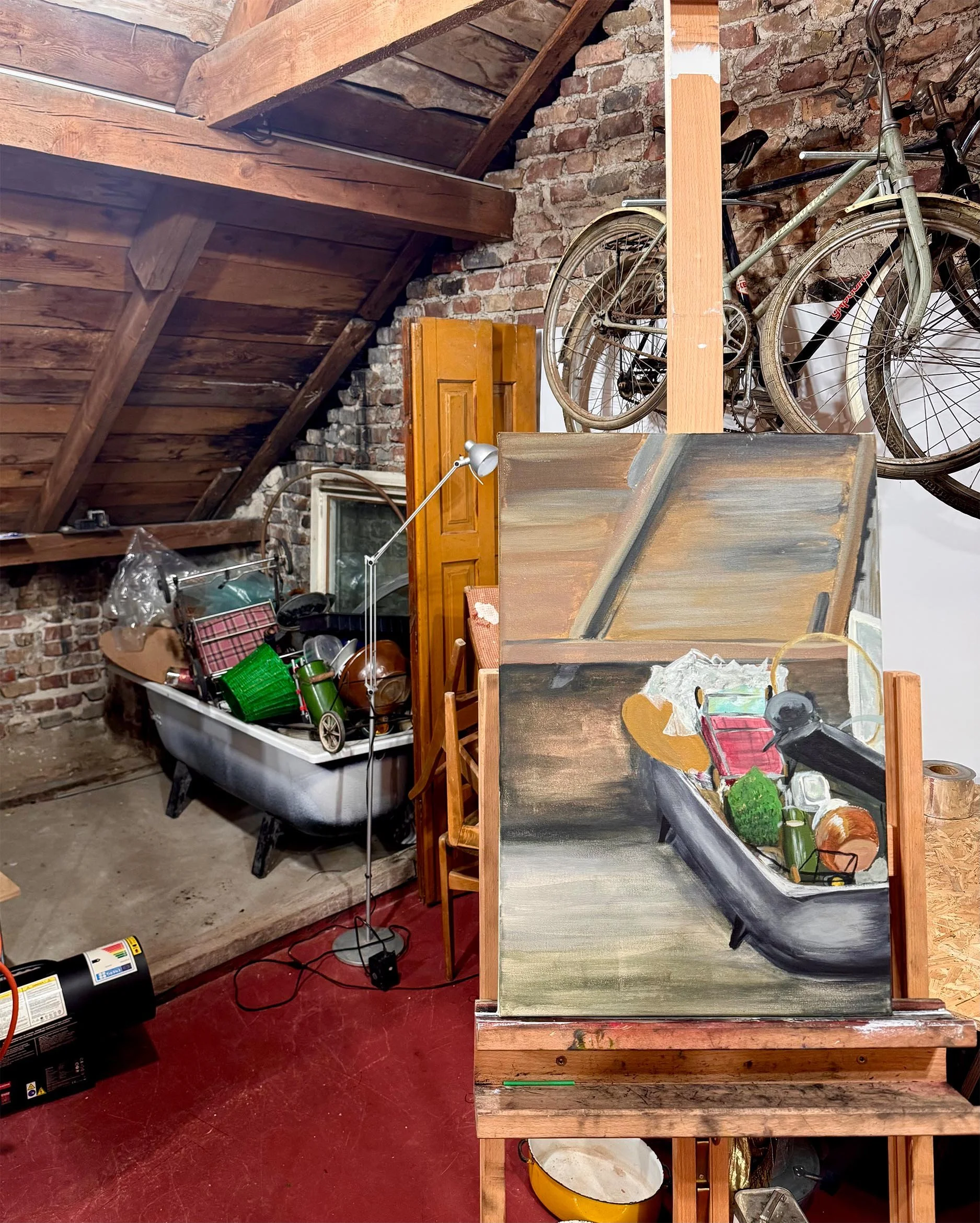 A room with wooden ceiling beams, brick walls, and a red floor. The room contains a bathtub filled with various household items, a floor lamp, a muskie painting, and exposed wooden furniture.
