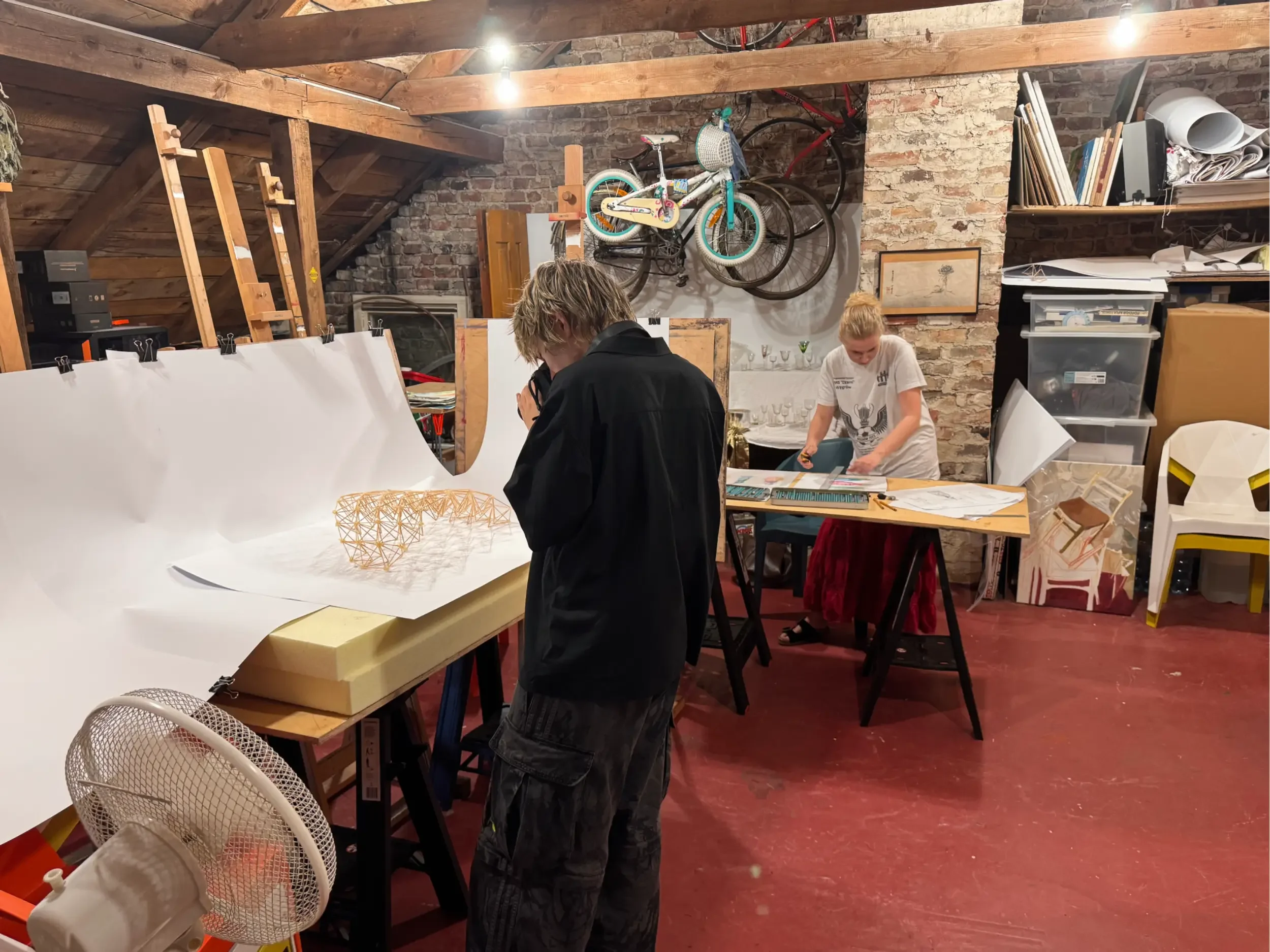 Dwie osoby pracują w pracowni artystycznej Mińsk Mazowiecki. Mężczyzna na pierwszym planie robi zdjęcie swojej strukturze z patyczków. Ustawił ją na tle białego kartonu. Na drugim planie kobieta stoi i rysuje kredkami projekt scenografii do teczki.