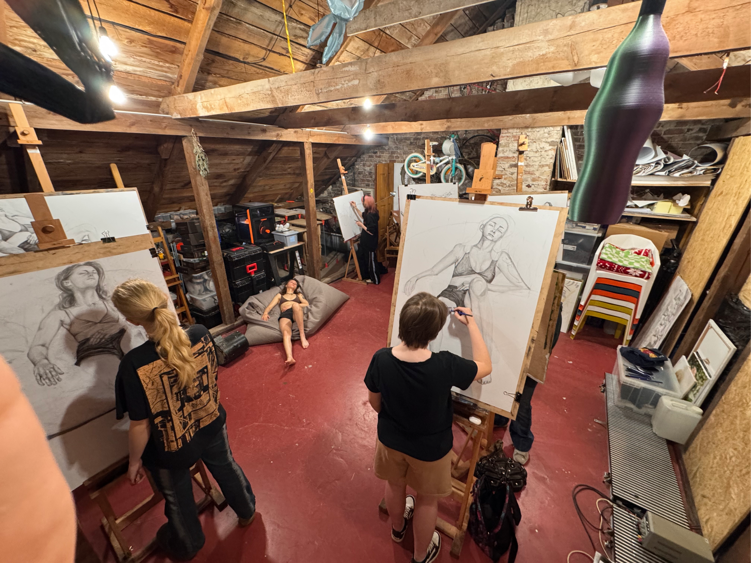 An art studio in an attic with several people drawing and painting. There are large unfinished portraits of women on easels, a woman lying on a couch, and others standing and working on their art. The room has wooden beams, shelves with art supplies,