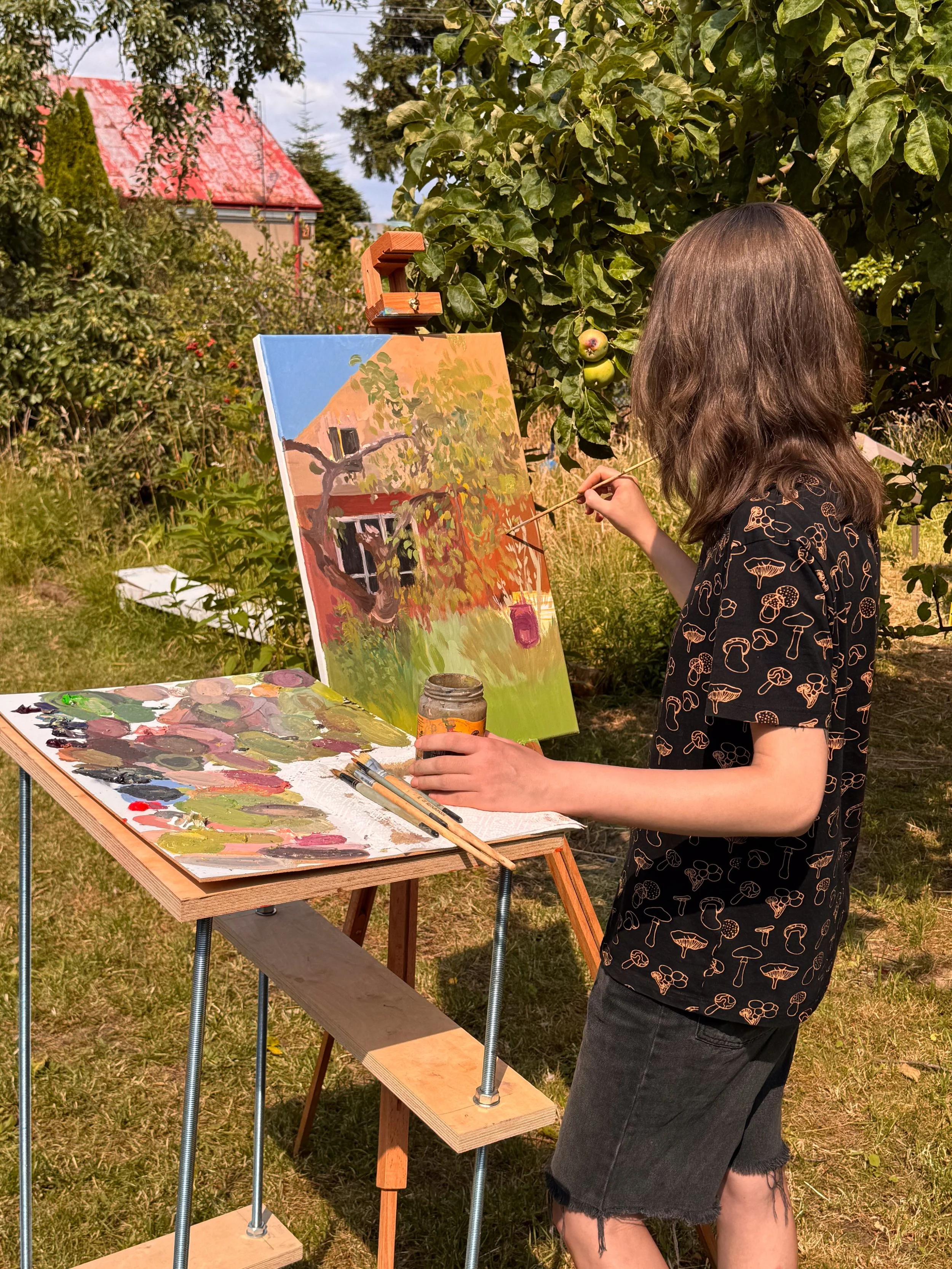 A person with brown hair and wearing a black shirt with mushroom patterns is painting outdoors. The artist is standing next to an easel with a canvas depicting a house with orange and green foliage. There are painting supplies on a table nearby, surrounded by trees and a red-roofed building in the background.
