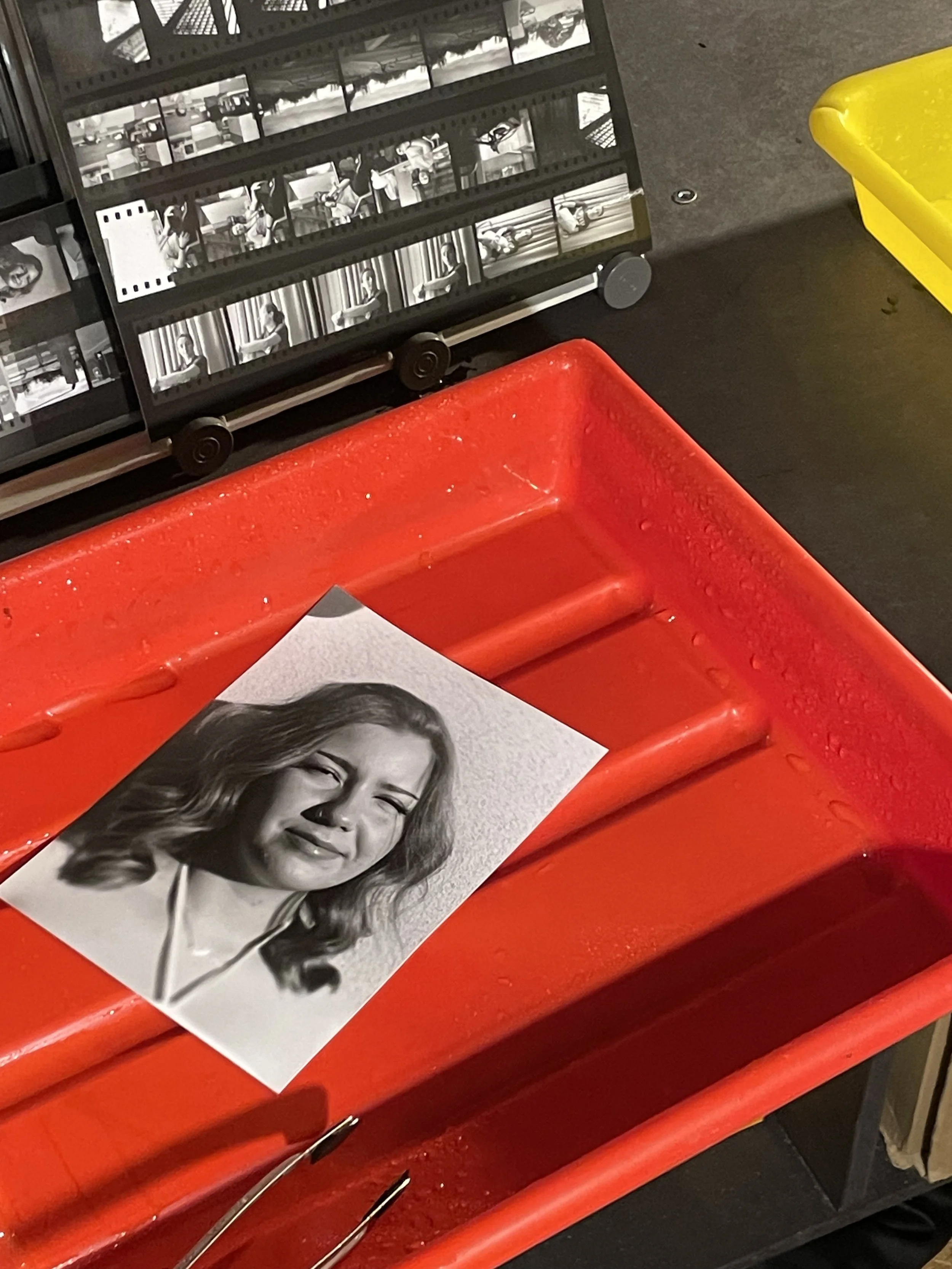 A black and white photograph of a woman smiling, printed on a piece of paper placed on a red chair. In the background, there is a film strip with multiple scenes in black and white.