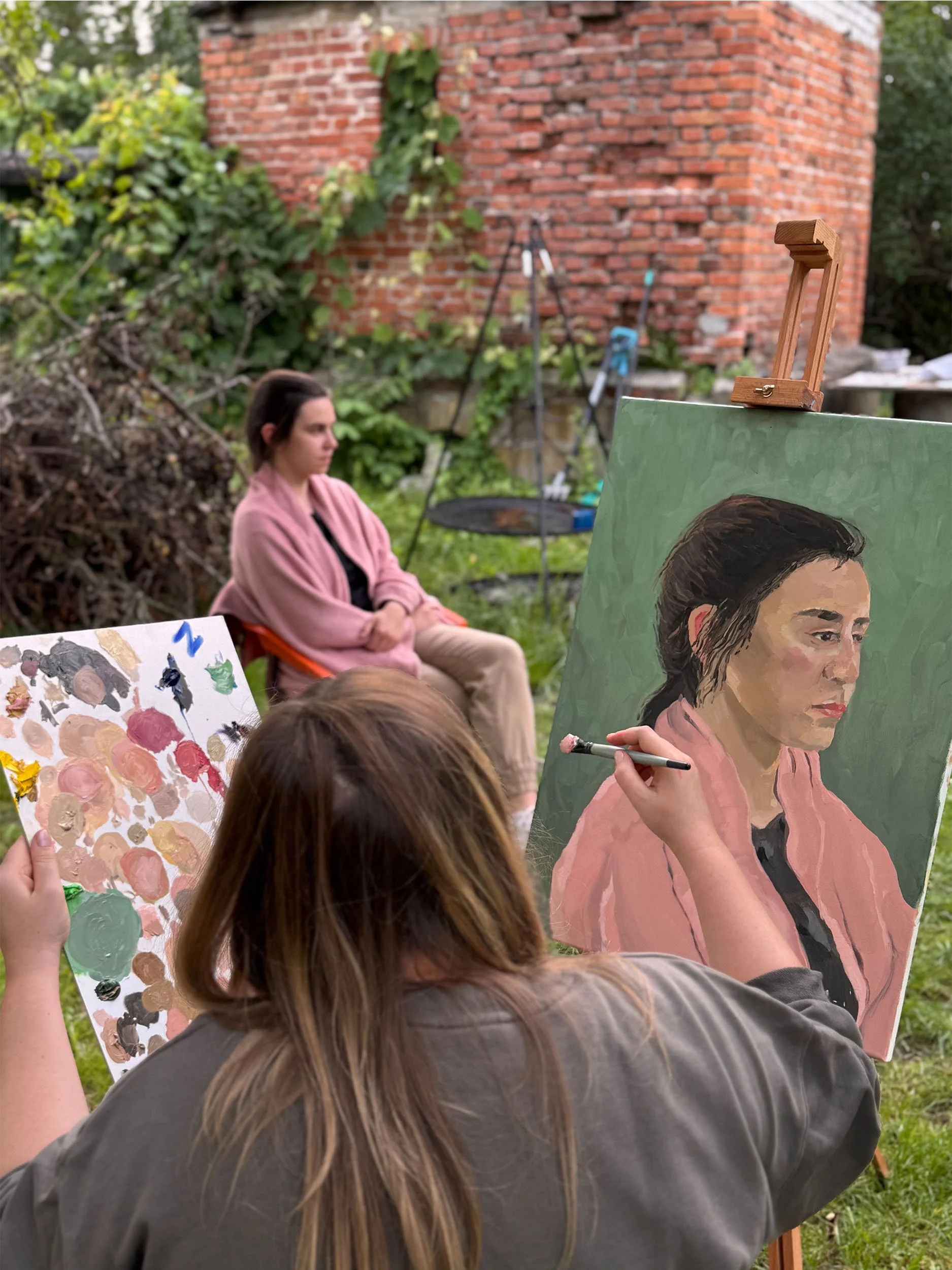 Multiple people, including children, are outdoors at an art event. One person with long brown hair is painting a portrait of a woman with dark hair, wearing a pink jacket, on a canvas. Another person is holding a palette with various paint colors. Th