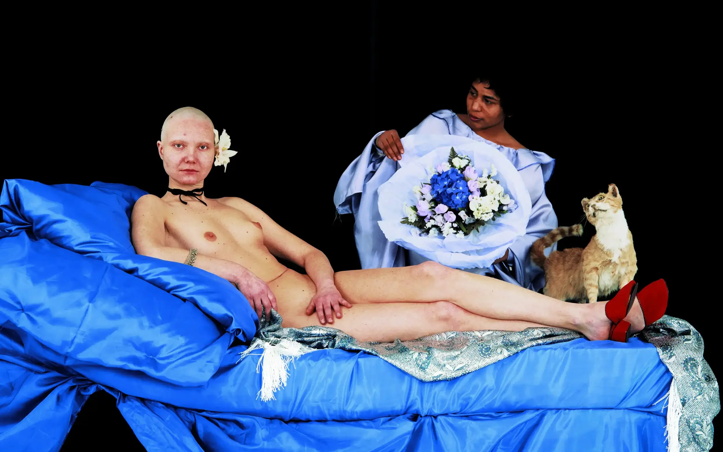 A person with a shaved head and a flower in their ear lying on a blue silk sheet, with another person holding a bouquet of flowers and a cat sitting nearby against a black background.