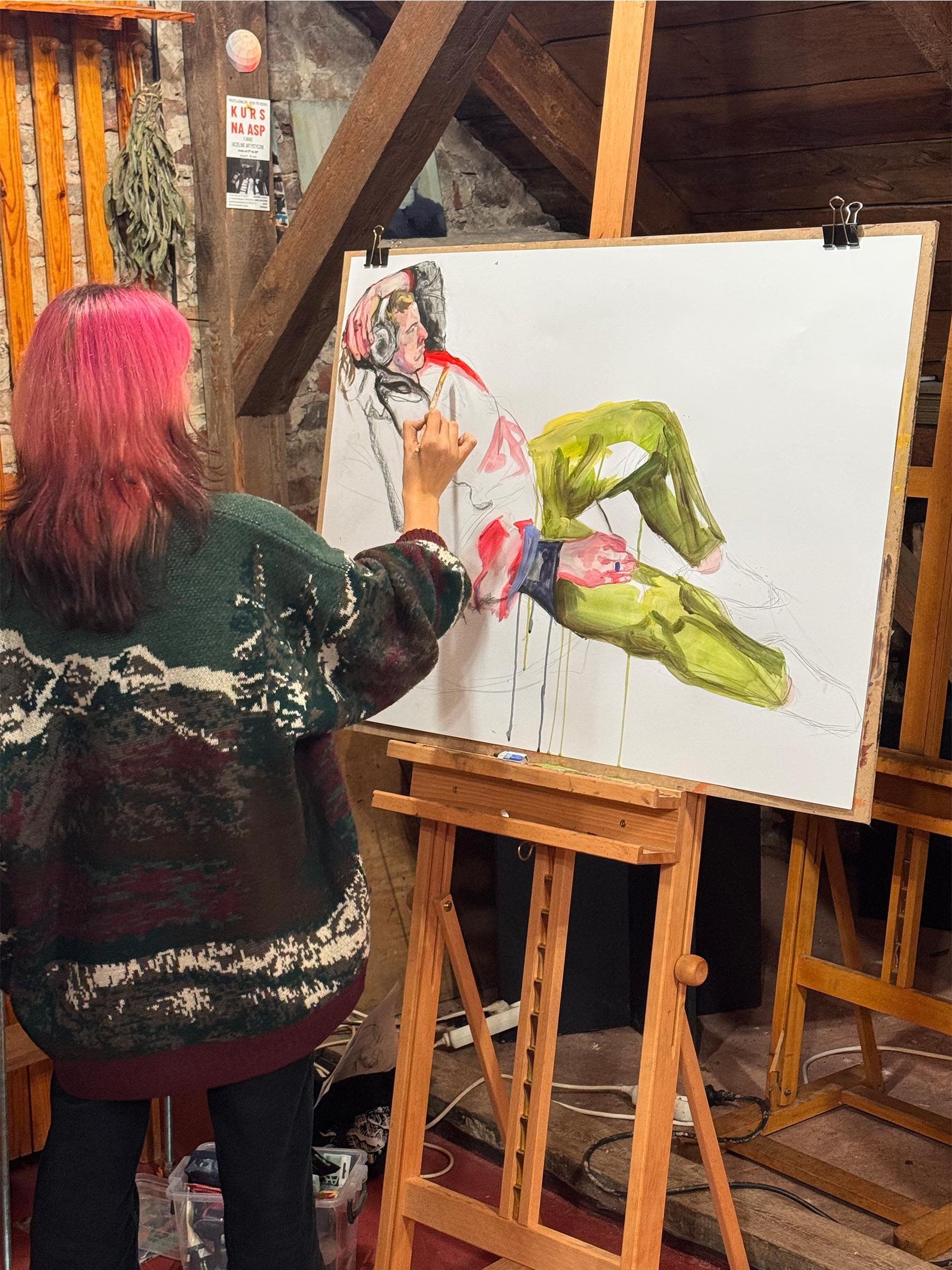 A woman with pink hair paints a realistic portrait of a woman sitting with her legs crossed and wearing headphones, on a large canvas in an art studio with wooden beams and stone walls.