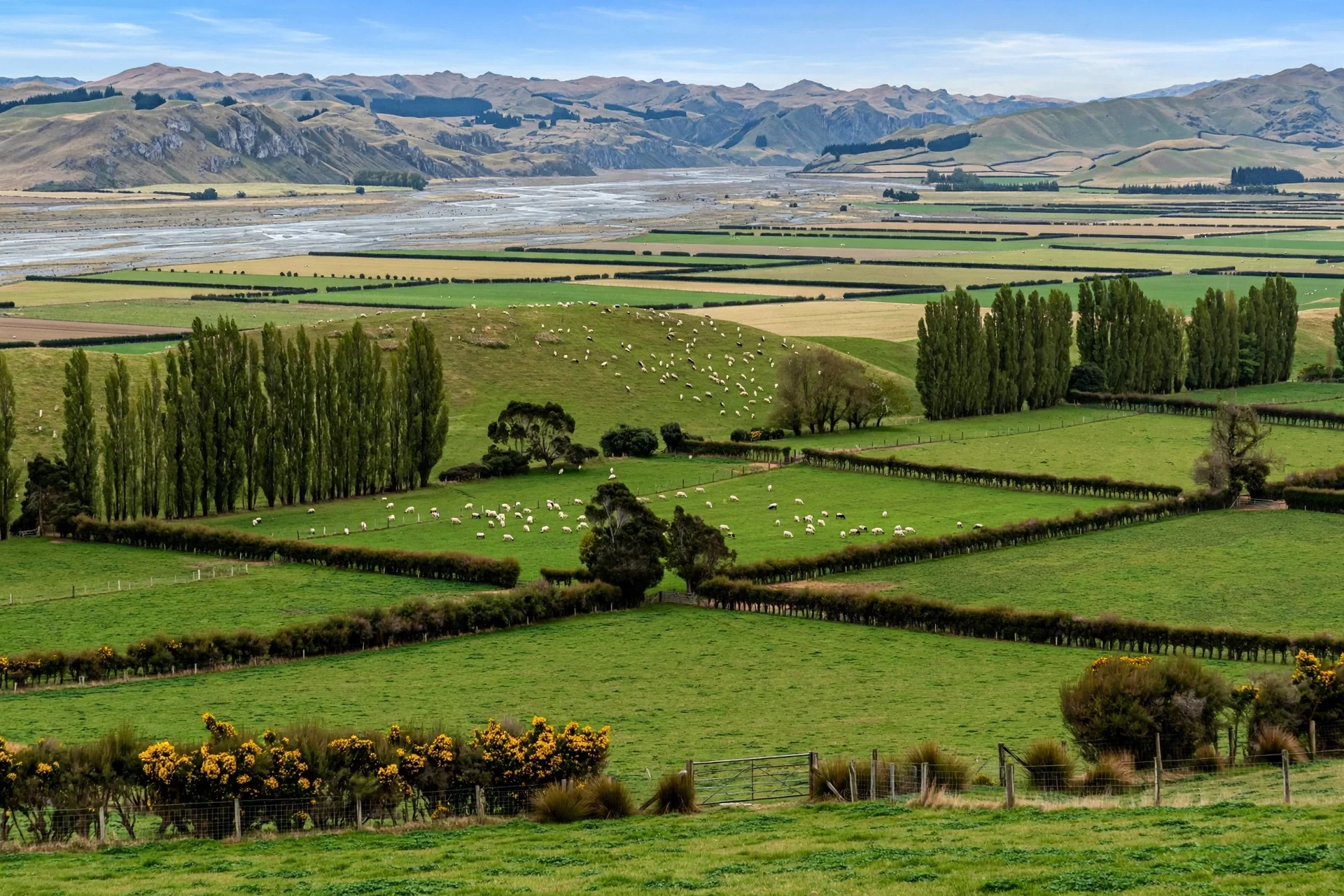 Scenic farmland with rolling green fields, tall trees, sheep grazing, and distant mountains under a partly cloudy sky.