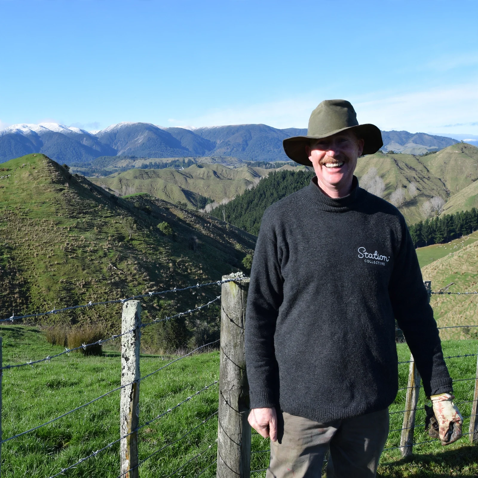 A smiling man wearing a wide-brimmed hat and a black sweater with the logo 'Station Collective' standing outdoors near a fence post in a scenic mountainous landscape with green hills and snow-capped peaks in the background.