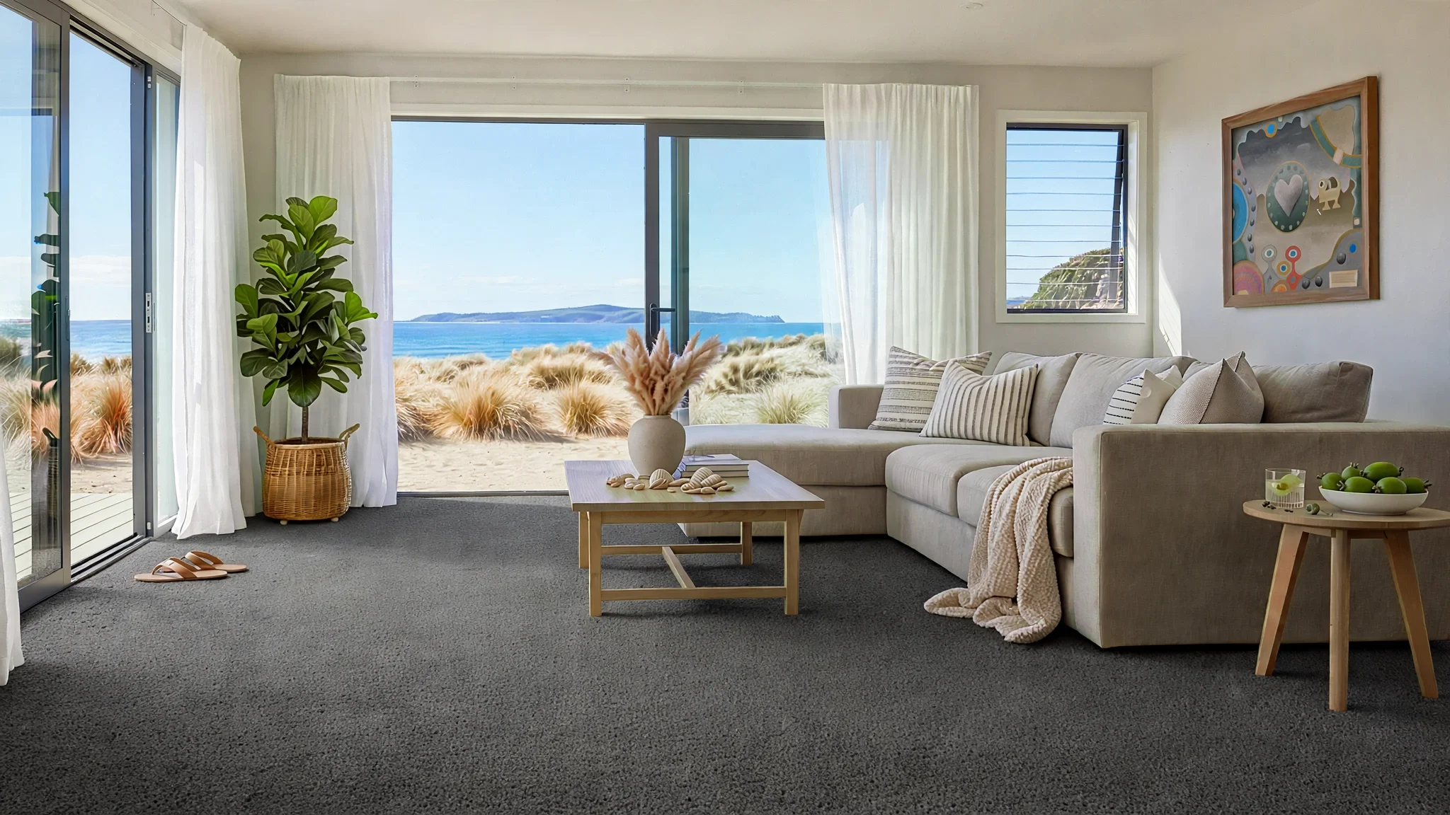 Living room with beige sectional sofa, wooden coffee table with seashell decor, potted plant, curtains, ocean view through large glass sliding door and window, abstract art on wall, small side table with green apples, and sandy beach outside.