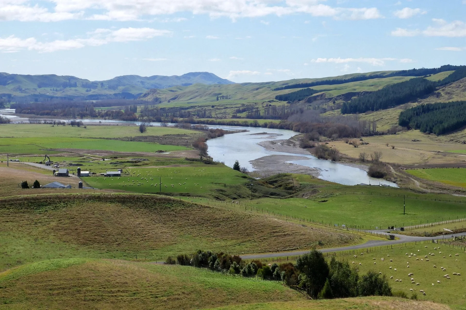 A scenic landscape of rolling green hills with a river winding through the valley, scattered trees, farm buildings, and distant mountains under a partly cloudy sky.