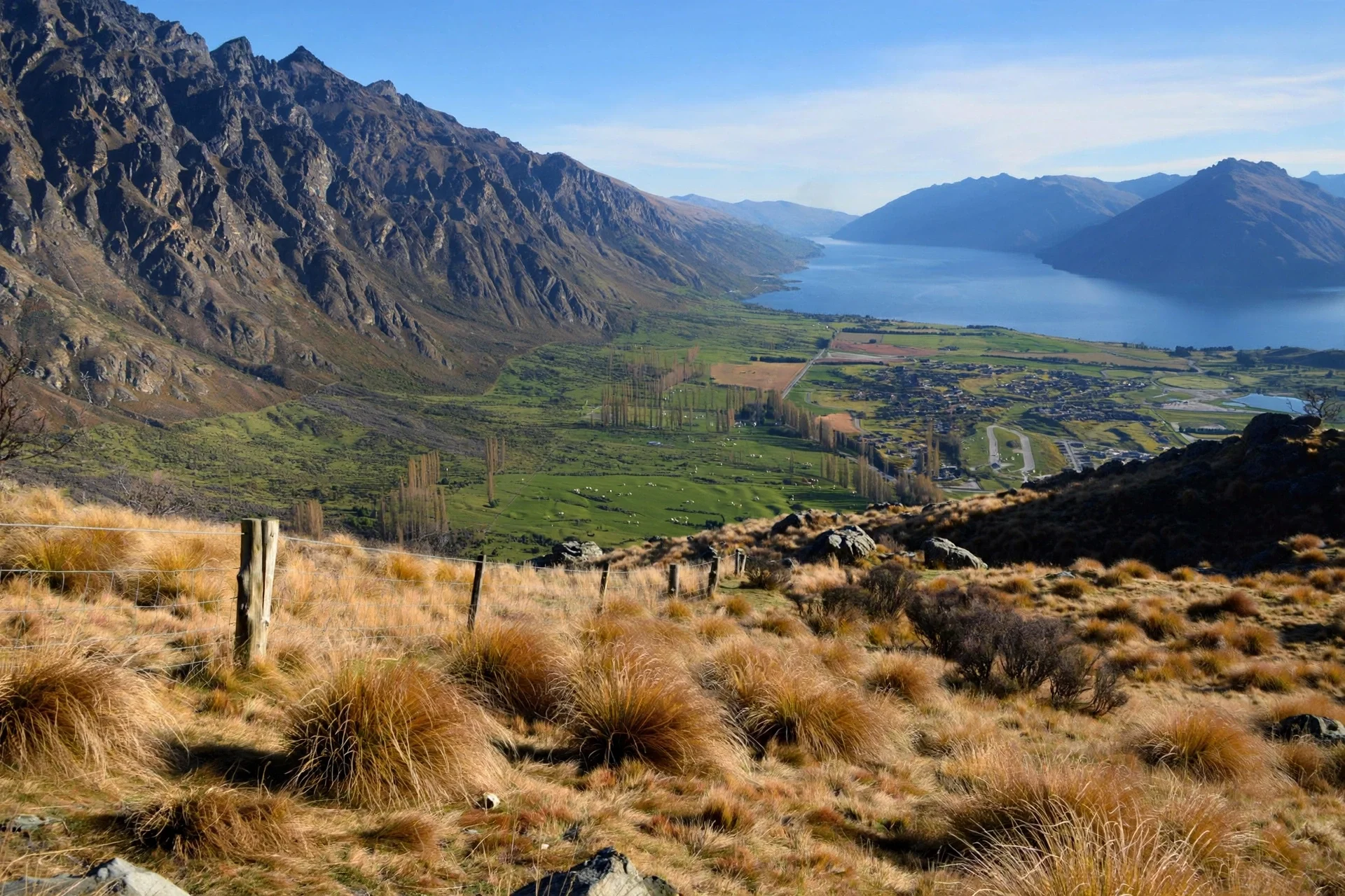 Scenic landscape with rugged mountains on the left, a wide lake in the middle, and a lush green valley below with small buildings and winding roads, under a clear blue sky.