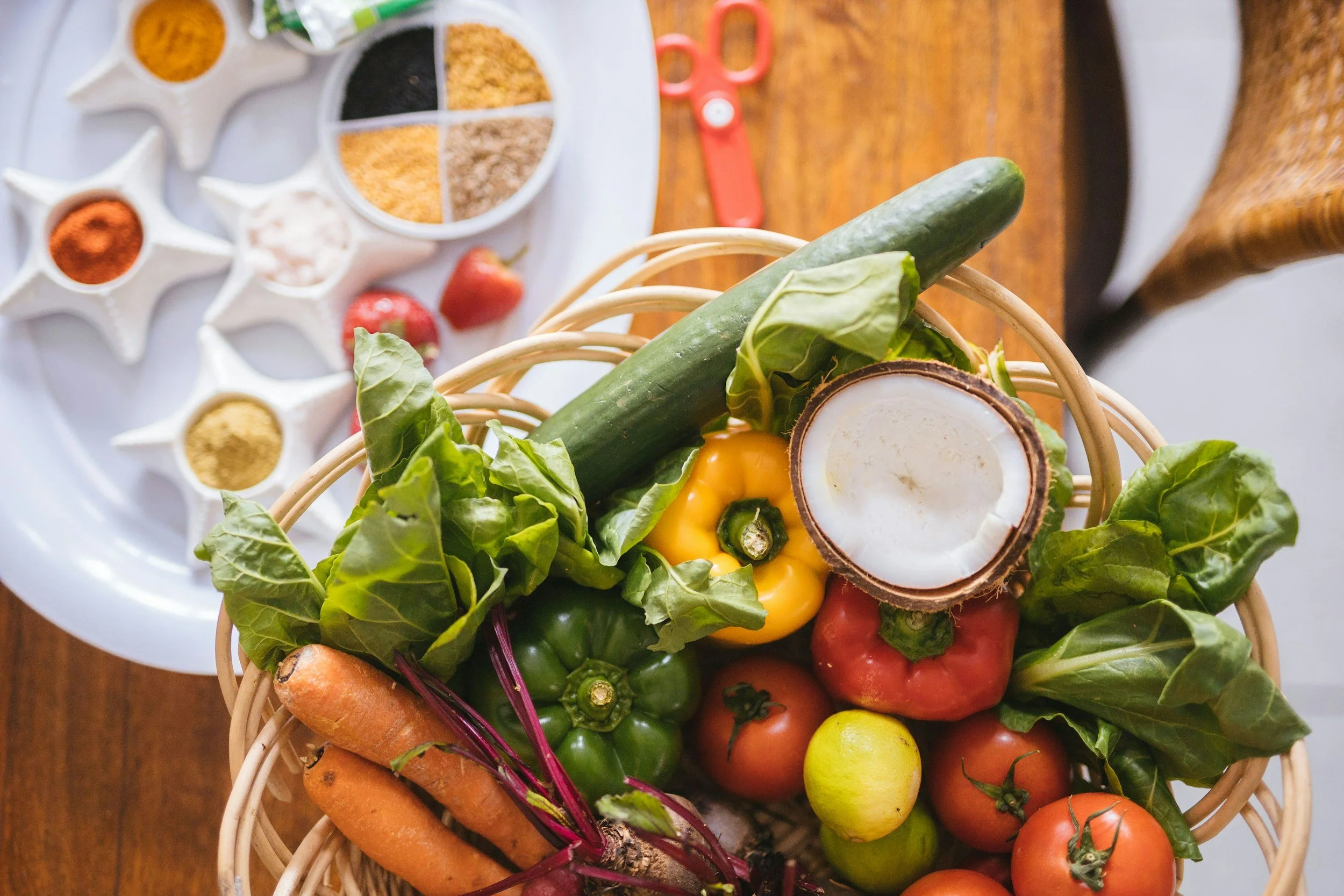 A basket filled with fresh vegetables including carrots, yellow bell peppers, green and red tomatoes, a cactus pear, and leafy greens on a wooden surface. In the background, there is a white tray containing strawberries and a variety of spices in small white dishes, along with a pair of red scissors.