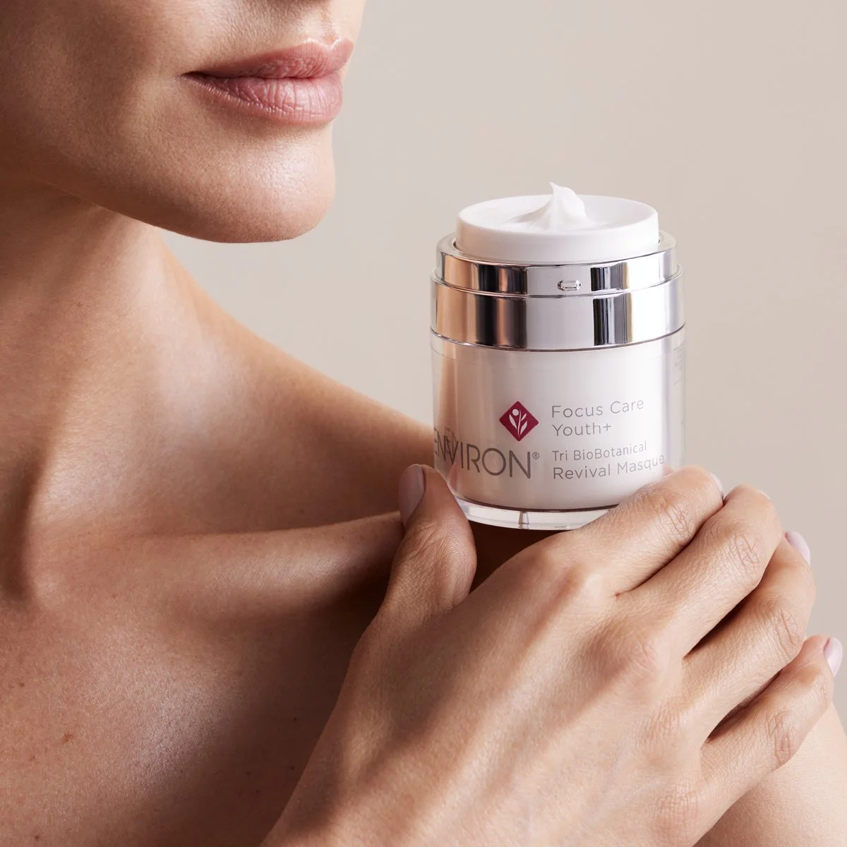 A woman holds a jar of Environ Focus Care Youth+ Tri BioBotanical Revival Masque near her shoulder. The jar is open, revealing a white cream inside.