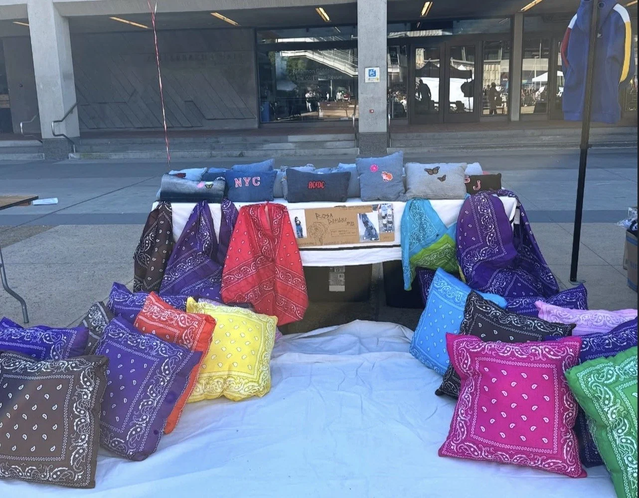 A street vendor display of colorful bandanas and small embroidered pillows, with a downtown city background behind glass doors.