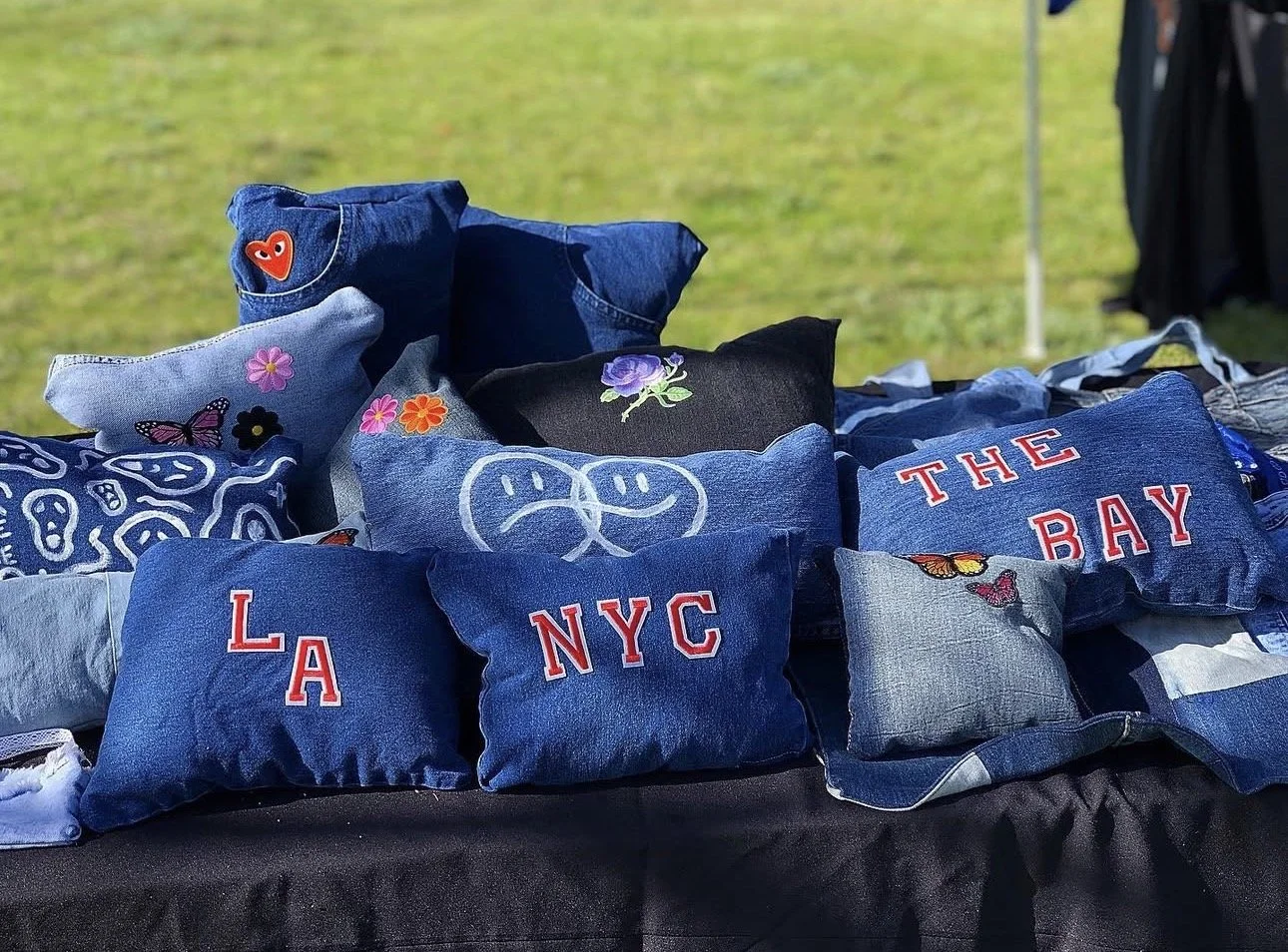 A display of small denim and fabric pouches with various embroidered designs, including words like 'LA,' 'NYC,' and 'THE BAY,' as well as floral and butterfly patterns, arranged on a black tablecloth outdoors.