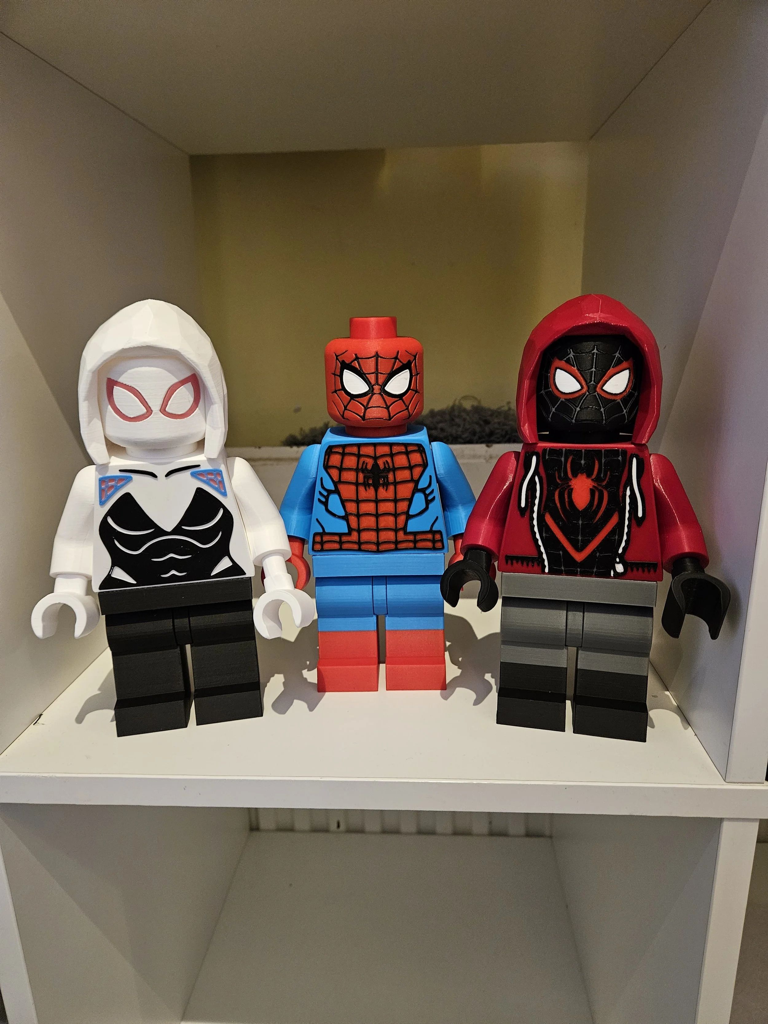 Three LEGO Spider-Man figures on a white shelf, each with a different costume and mask, face forward.