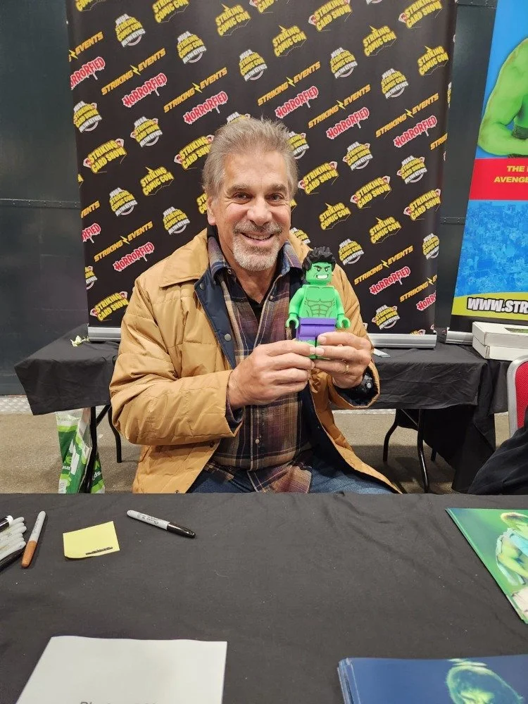 A man smiling and holding a green LEGO Hulk figure at a convention, with a backdrop featuring movie logos and a table with markers and papers.