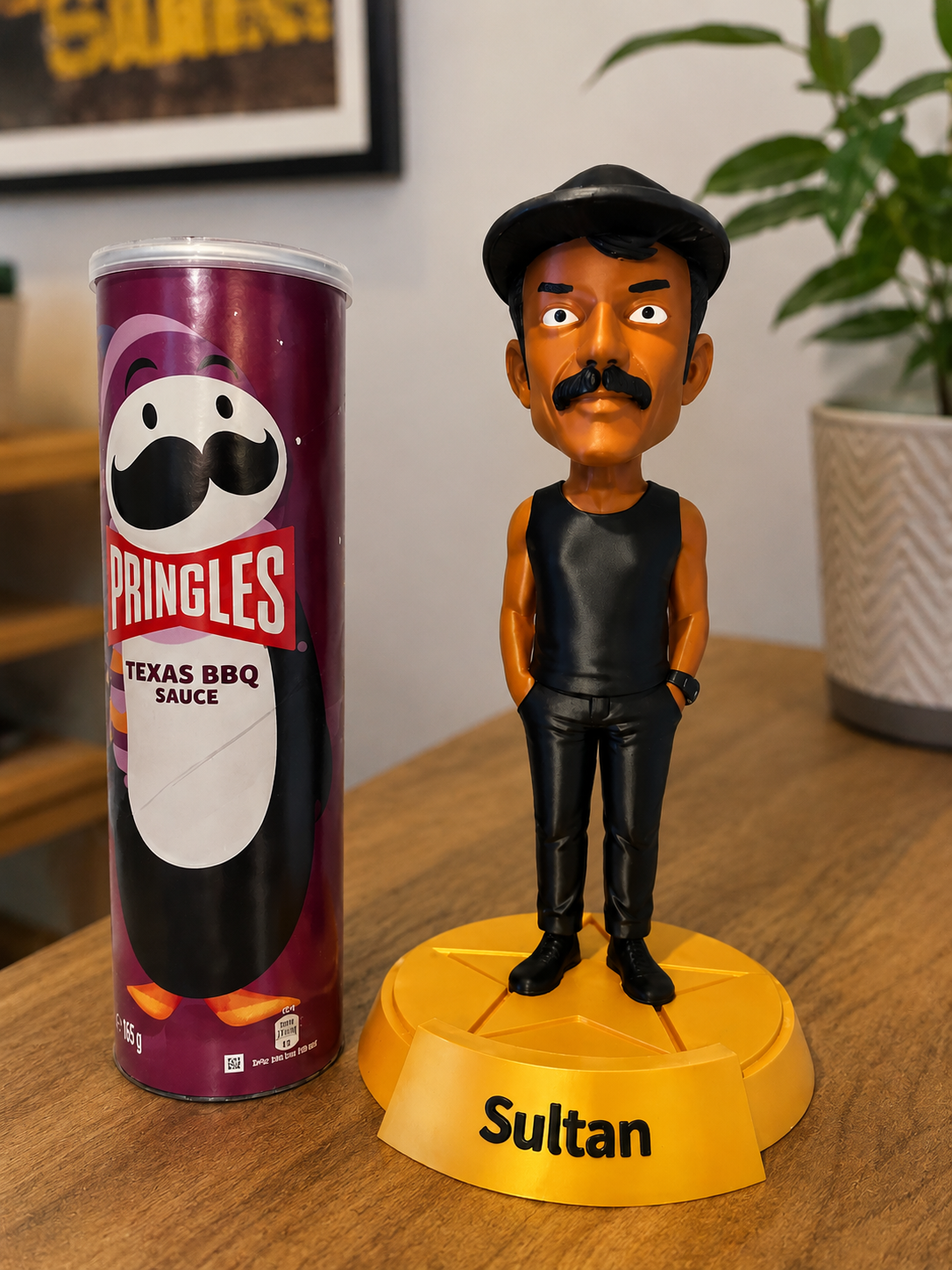 A Sultan bobblehead figure standing on a yellow base labeled 'Sultan' next to a can of Pringles Texas BBQ sauce on a wooden table, with a potted plant and wall decor in the background.