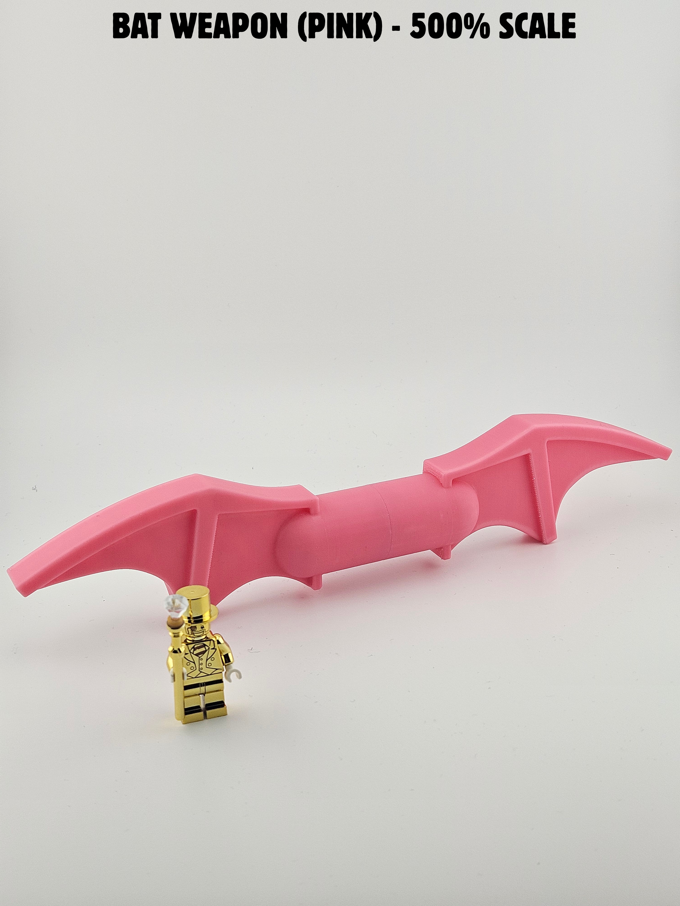 A pink plastic bat-shaped object with a small gold toy figure of a man in a tuxedo standing in front of it. The bat has architectural details and a small yellow accessory at one end. The background is plain white with text at the top reading 'Bat Weapon (Pink) - 50% Scale'.