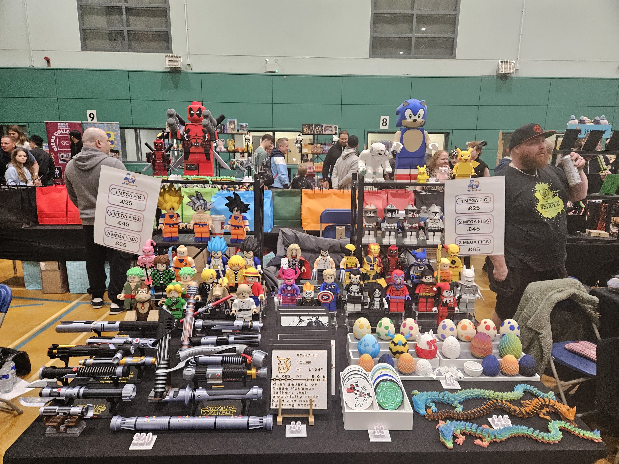 A toy convention booth displaying various LEGO characters, figures, and accessories, including popular characters like Deadpool, Sonic the Hedgehog, Pikachu, and Star Wars stormtroopers, with some pricing signs visible and people browsing and chattin