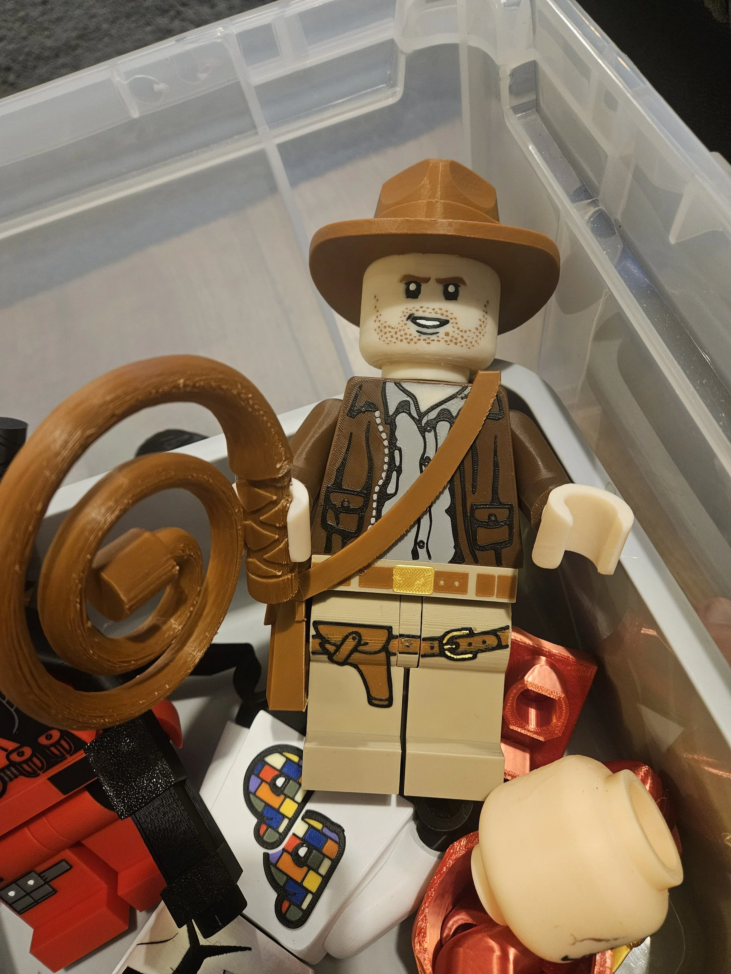 Lego toy figure resembling Indiana Jones, wearing a brown hat, jacket, and khaki pants, holding a whip, inside a plastic container with other objects.