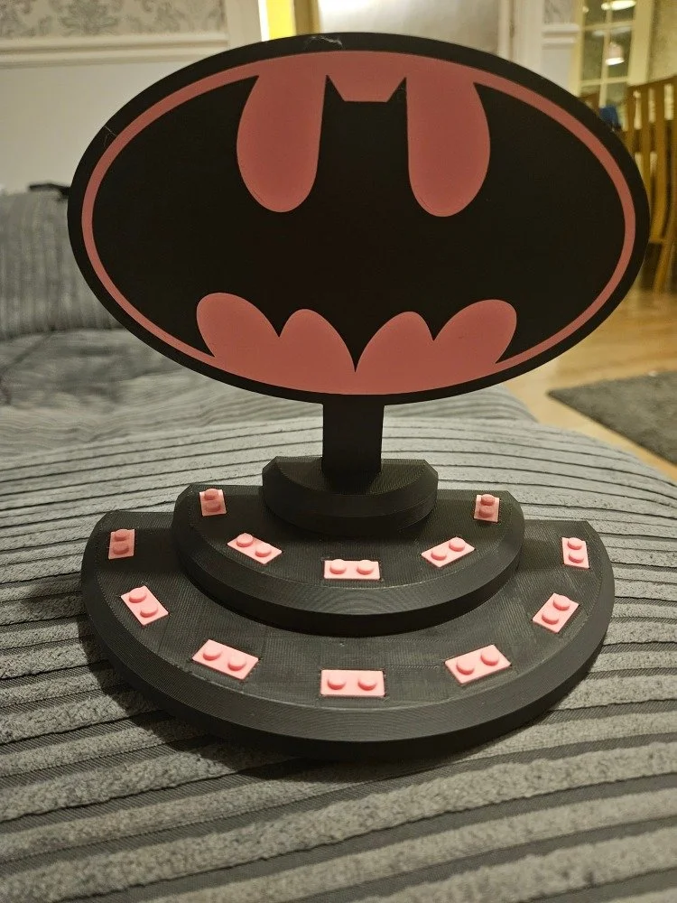 Lego Batman logo on a decorative stand with numbered pink Lego tiles, placed on a gray striped surface in a cozy home setting.