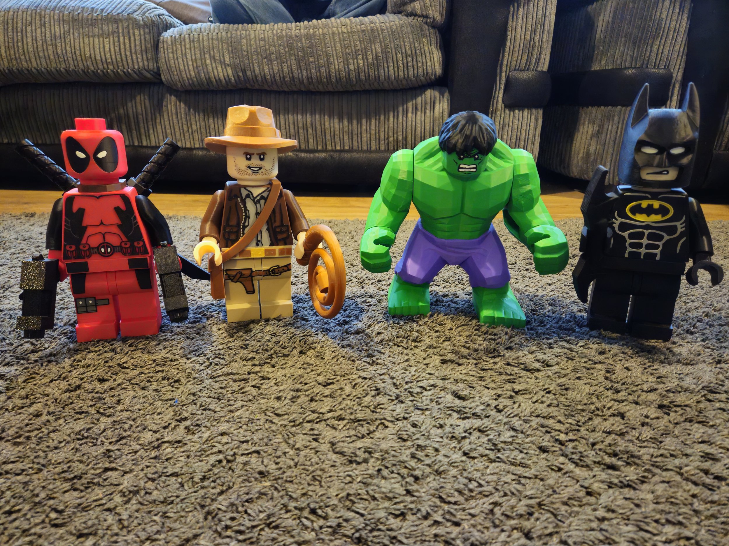 Photo of four LEGO superhero figures standing on a carpeted floor in front of a brown sofa. From left to right: Deadpool, Indiana Jones, Hulk, Batman.