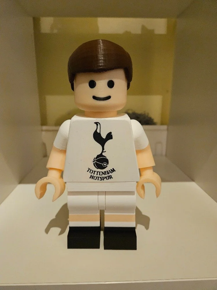Lego minifigure of a person with brown hair wearing a white Tottenham Hotspur football team jersey with the team's logo on it.