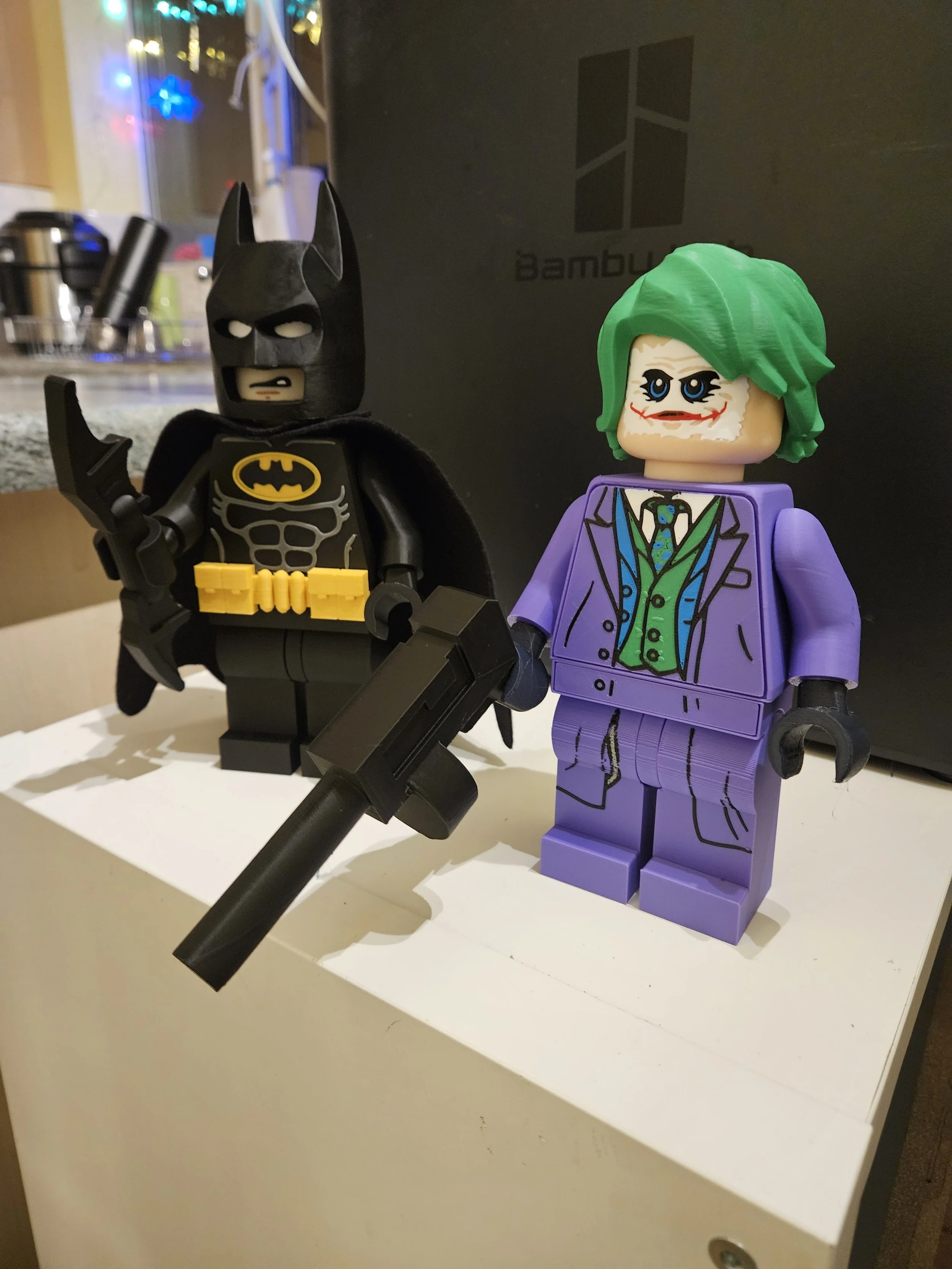 LEGO Batman and Joker figures on a white surface, with a black box and colorful lights in the background.