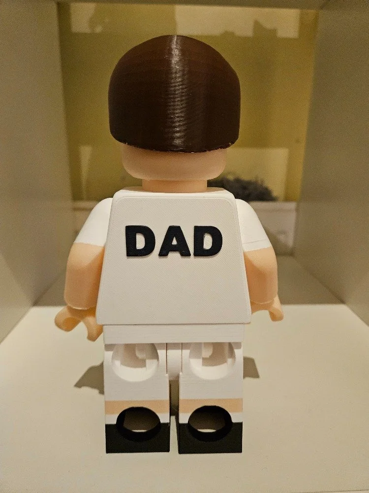 Back view of a Lego toy figure with brown hair, wearing a white shirt and white shorts, with a white vest labeled 'DAD' on the back, standing on a white surface.