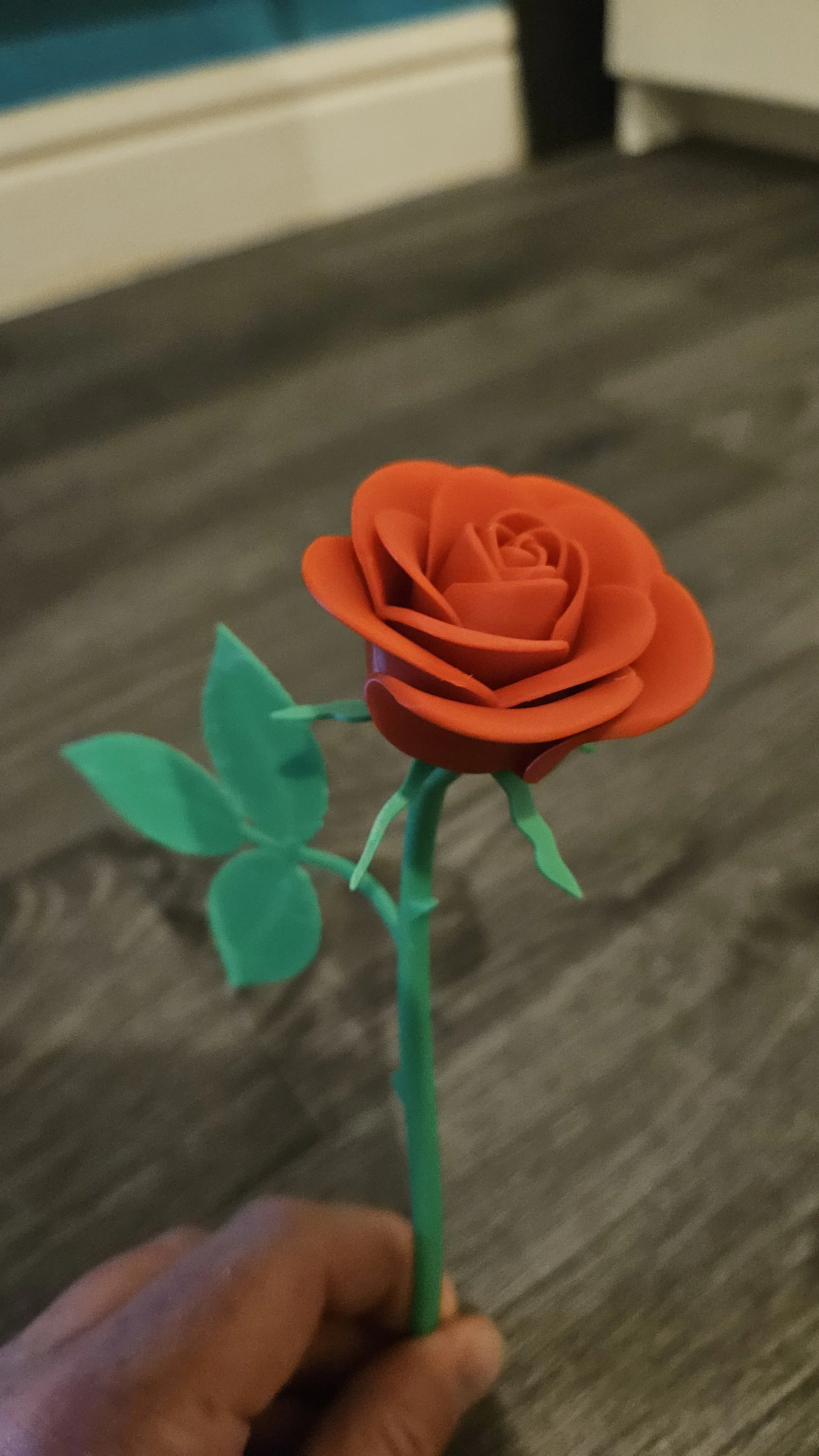 A plastic orange rose with green leaves on a green stem, held in a person's hand.
