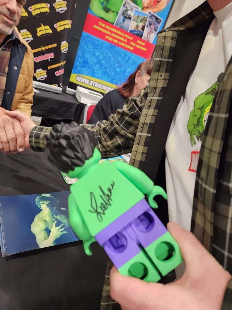 A person is holding a LEGO figurine with a signature on the back, in front of a table with glossy photographs and a man shaking hands with another person at an event or convention.
