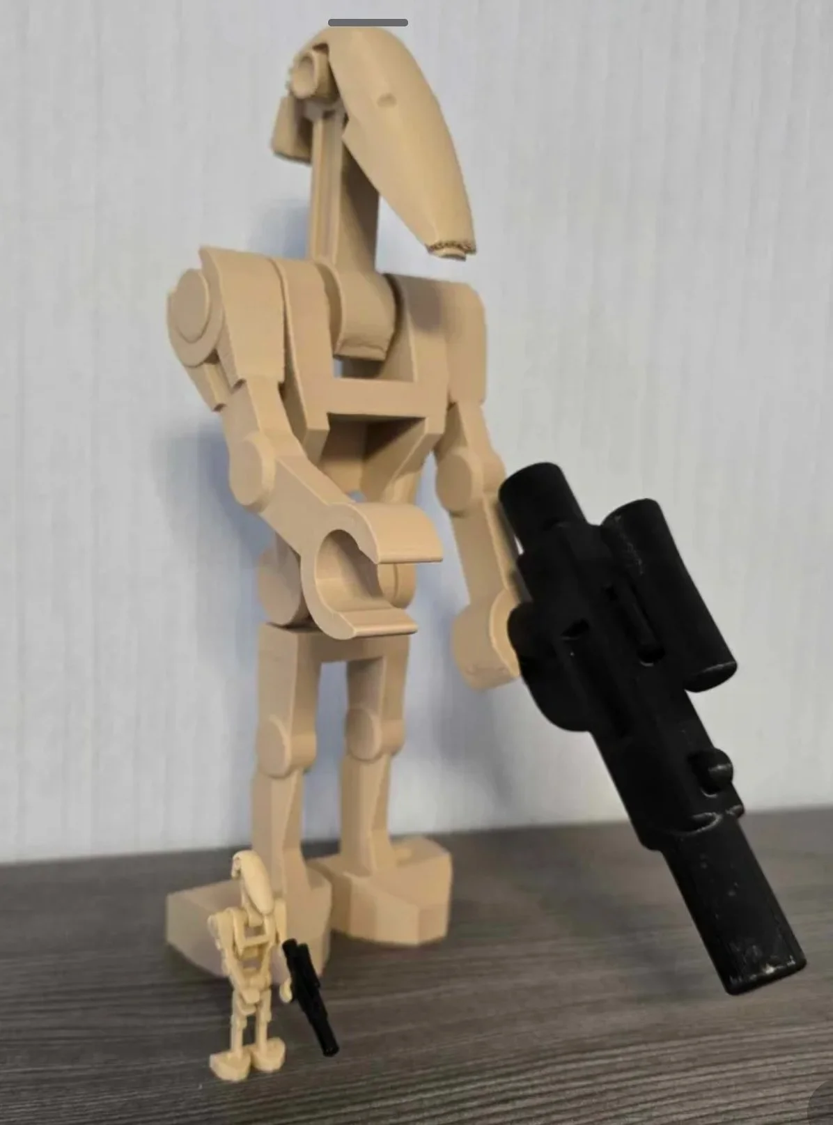 A wooden mannequin holding two black toy guns, one larger in its right hand and a smaller one in its left hand, standing against a plain white wall.