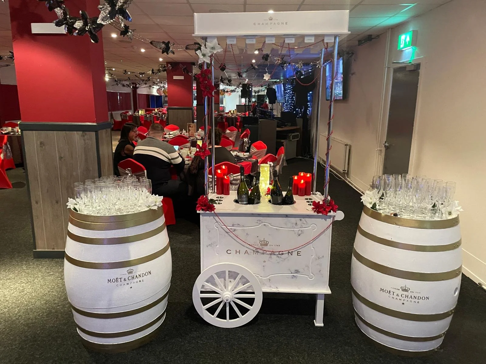 A festive champagne cart decorated with red flowers, candles, and champagne bottles, flanked by two large wine barrel displays with champagne glasses on top, in a decorated restaurant with tables set for a celebration.