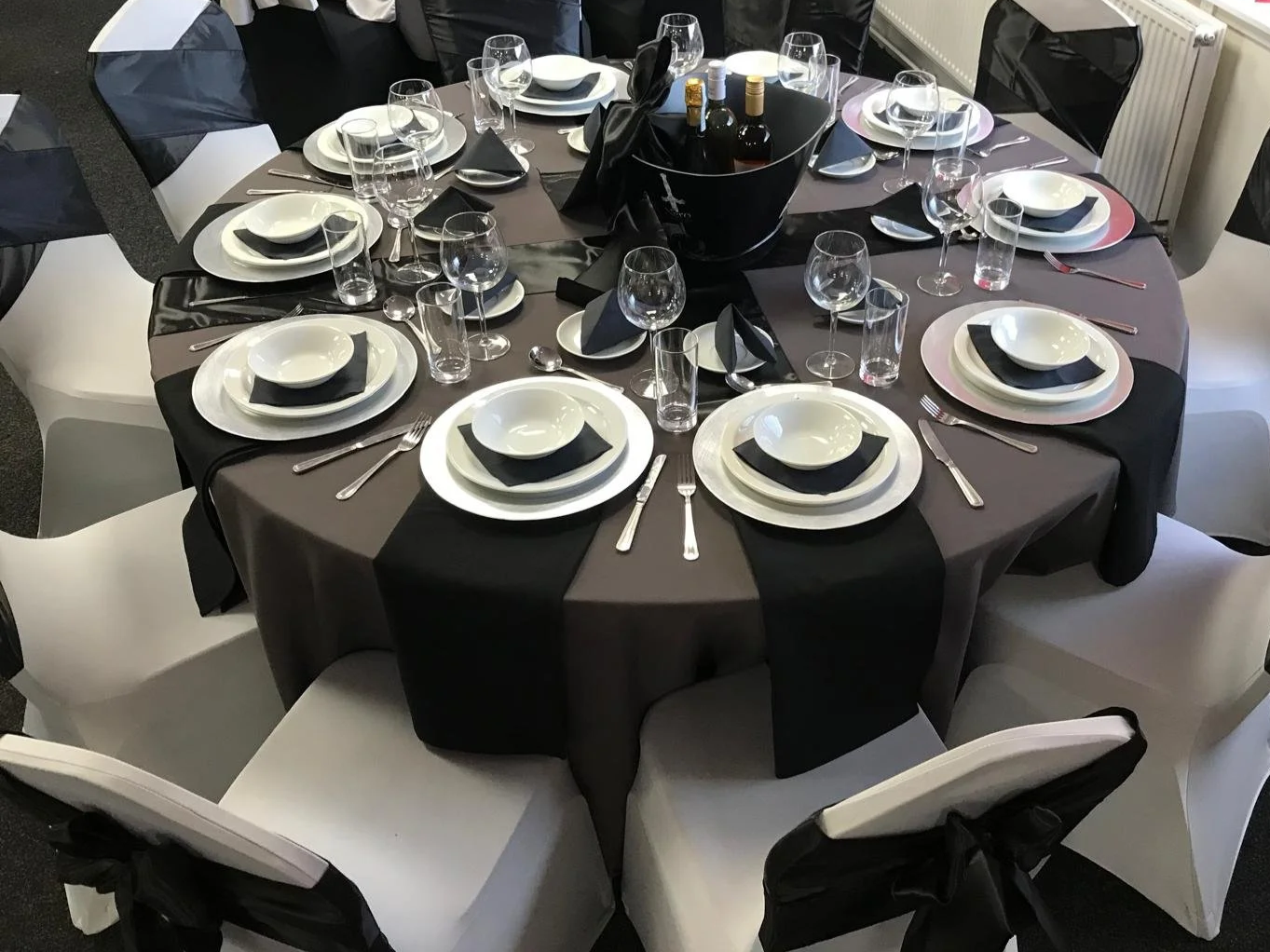 A round banquet table set for a formal event with black, gray, and white tableware, including plates, bowls, glasses, and silverware, with a centerpiece containing bottles of wine or champagne.