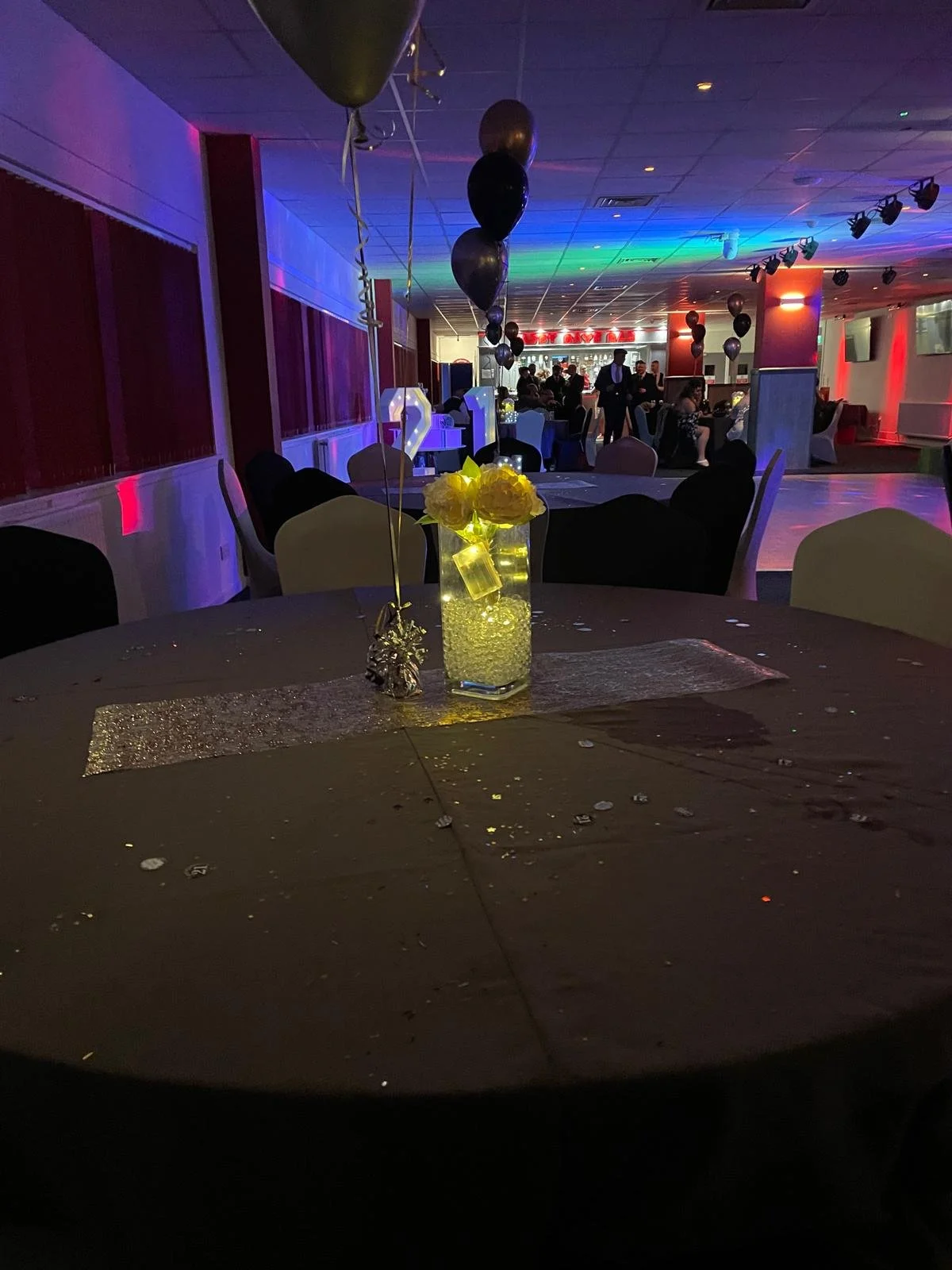 Decorated banquet table with a glowing candle centerpiece and black, silver, and gold balloons at a party or event, with people mingling in the background under colorful lighting.